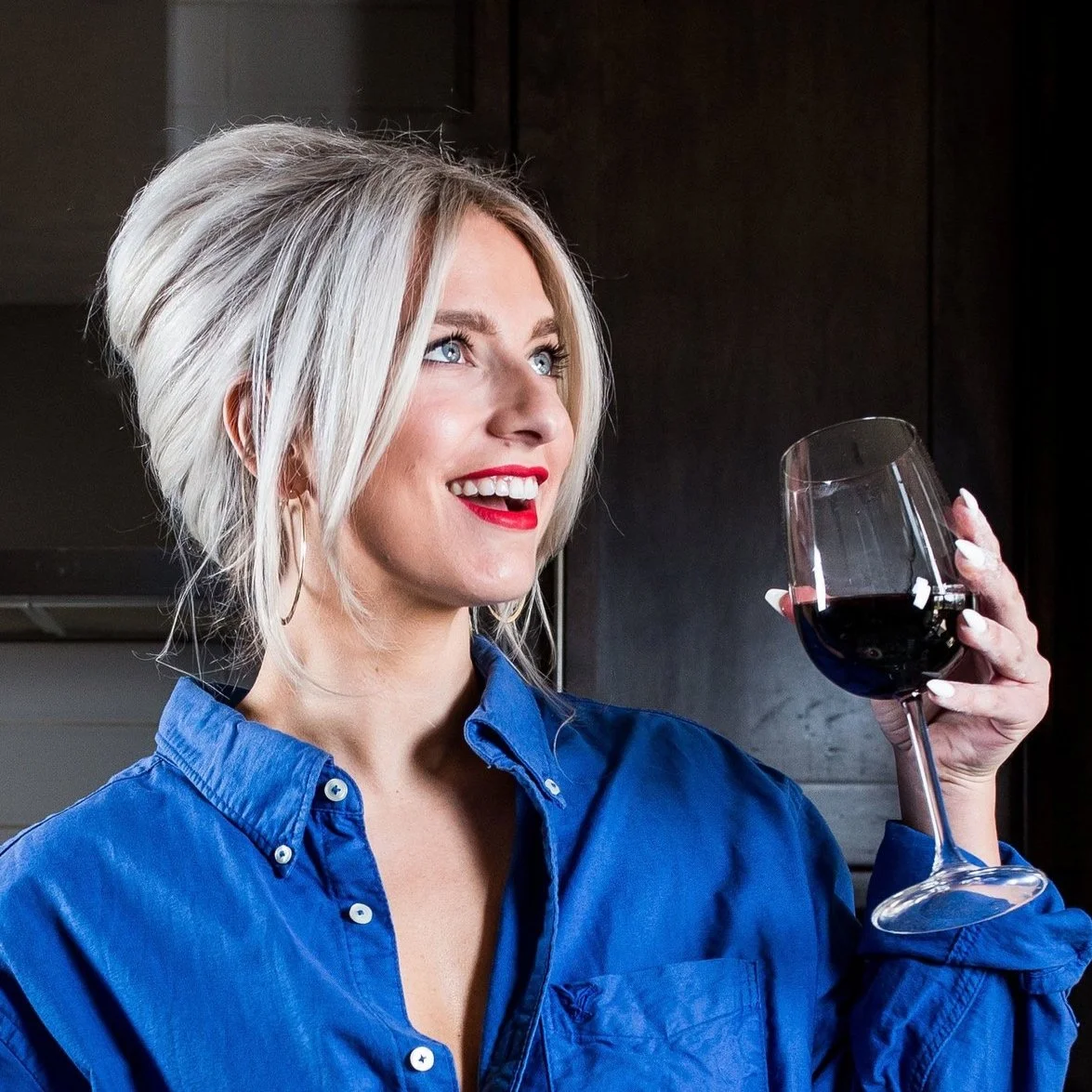 Breanna McDonald in a blue shirt holding a glass of red wine, smiling, with a dark background. Table Forty-Nine