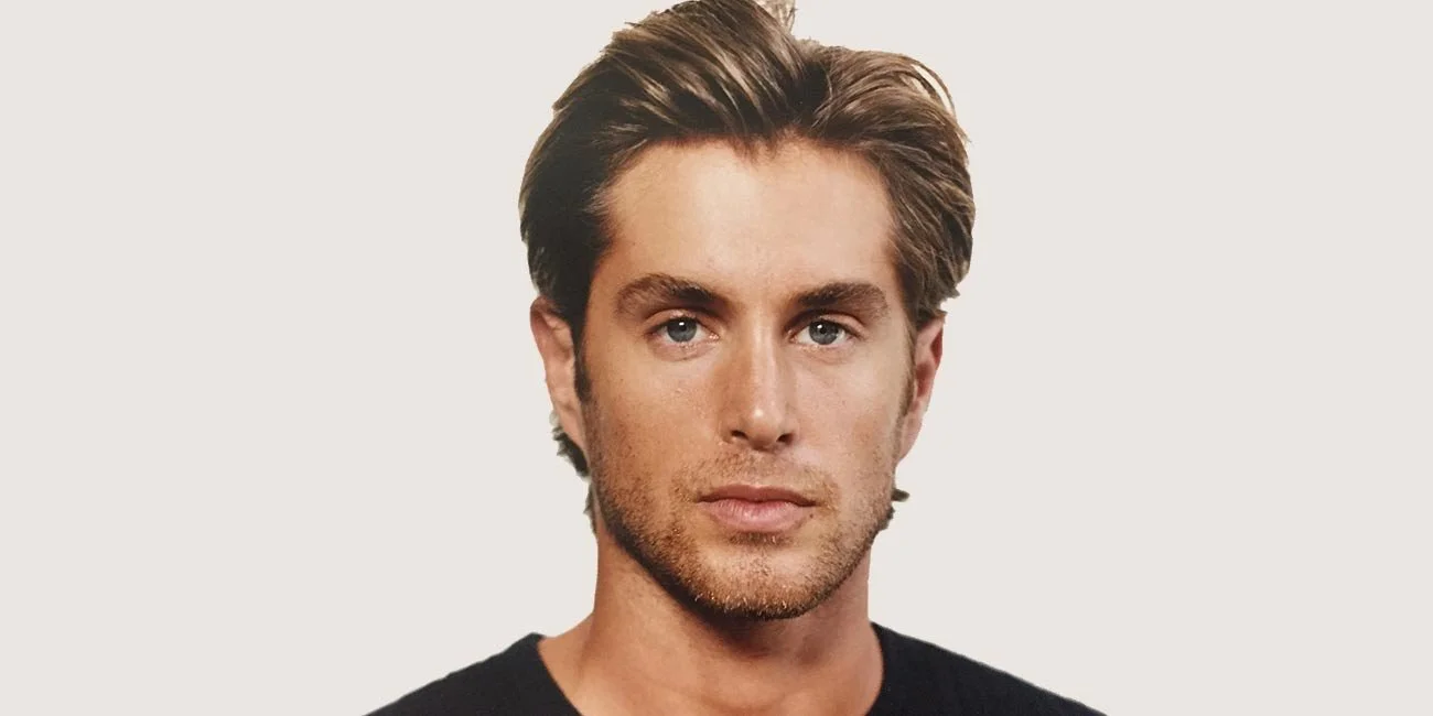 Portrait of Greg Sestero with styled hair and facial stubble, wearing a black shirt, on a neutral background.