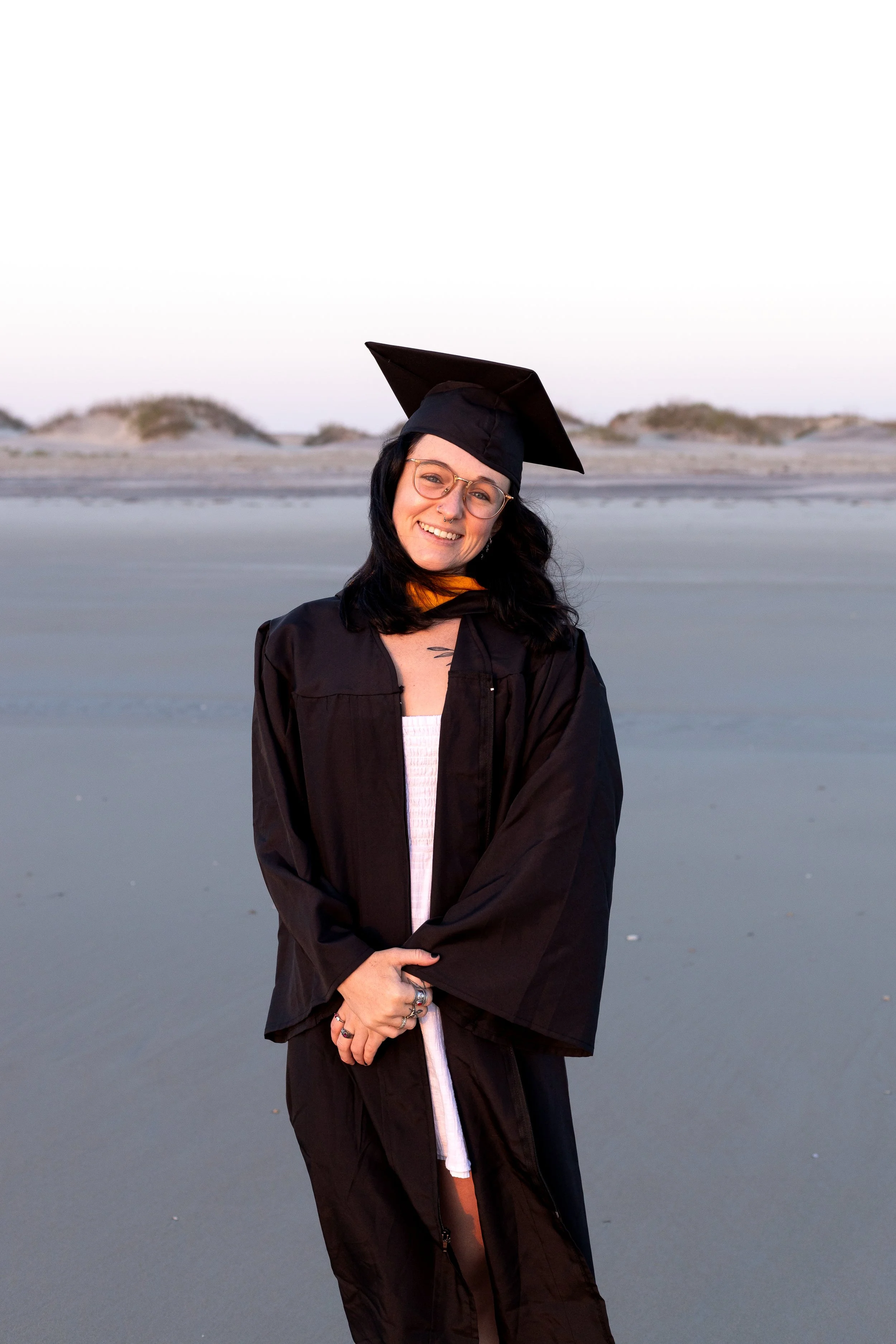 A young woman wearing graduation cap and gown, smiling, standing on a beach at sunset.