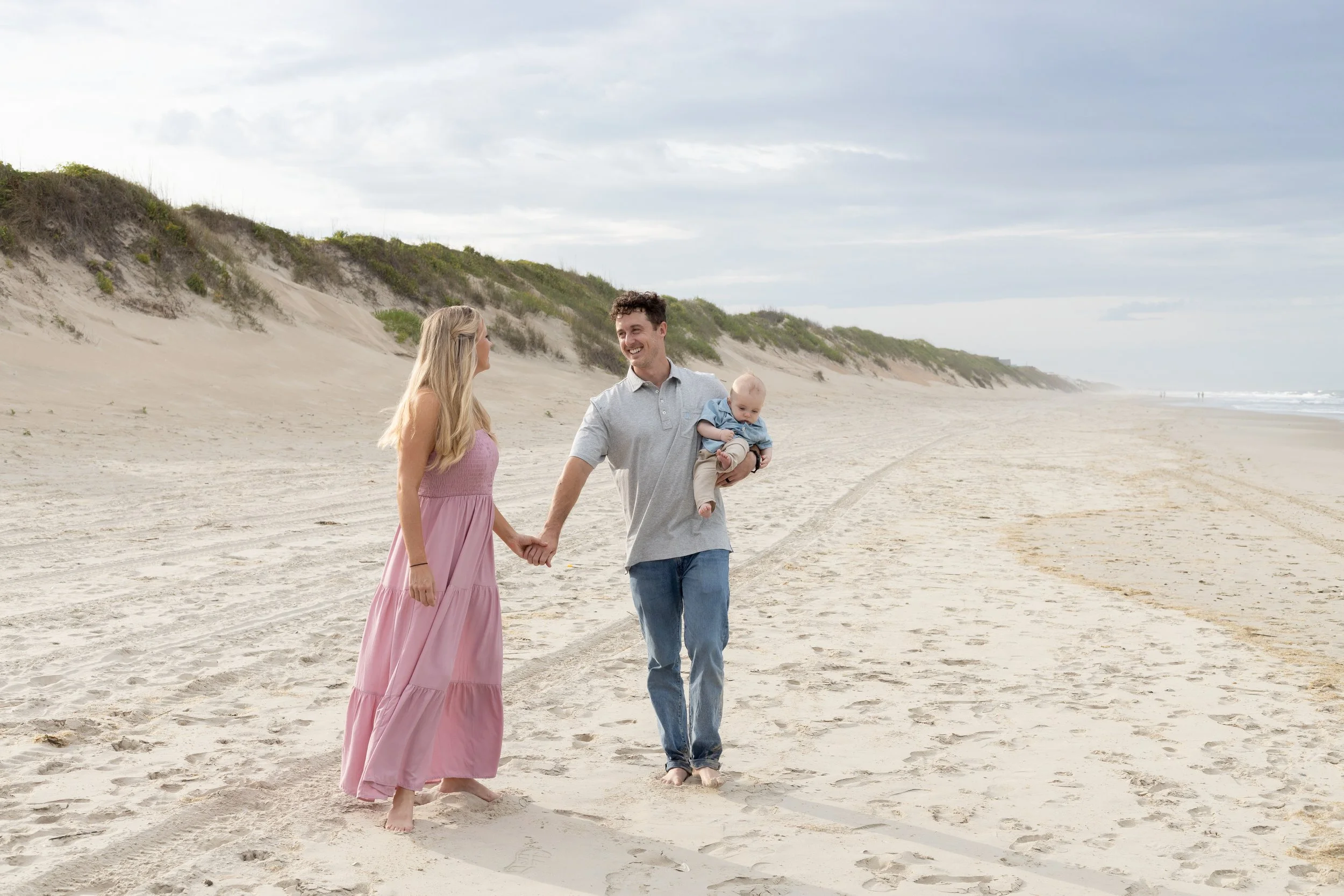 A happy family of three walking hand-in-hand on a sandy beach, with a woman in a pink dress, a man in a light gray shirt holding a baby, and a background of dunes and ocean under a cloudy sky.