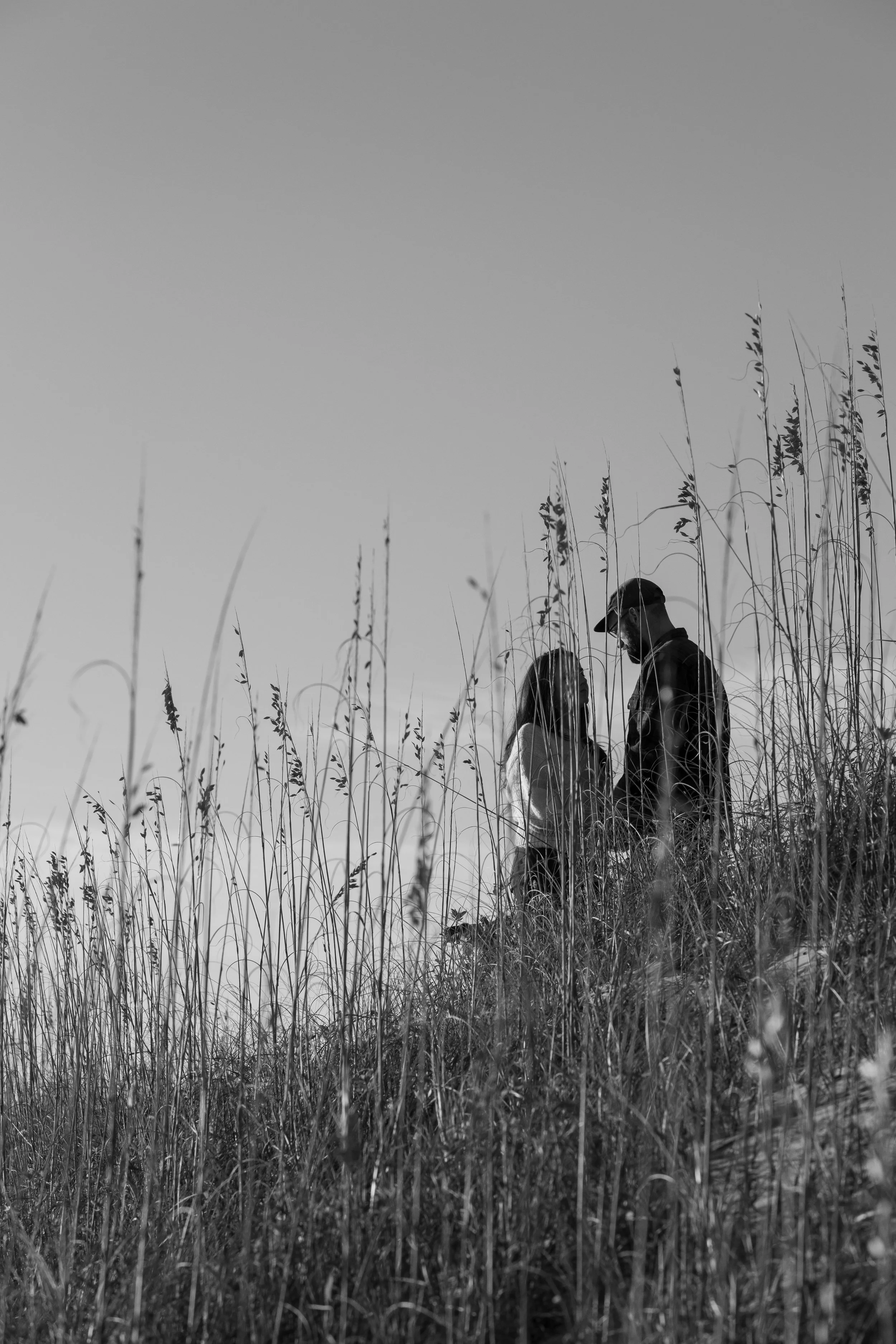 A black and white photo of a man and woman in a grassy field, facing each other and holding hands, with a clear sky in the background.