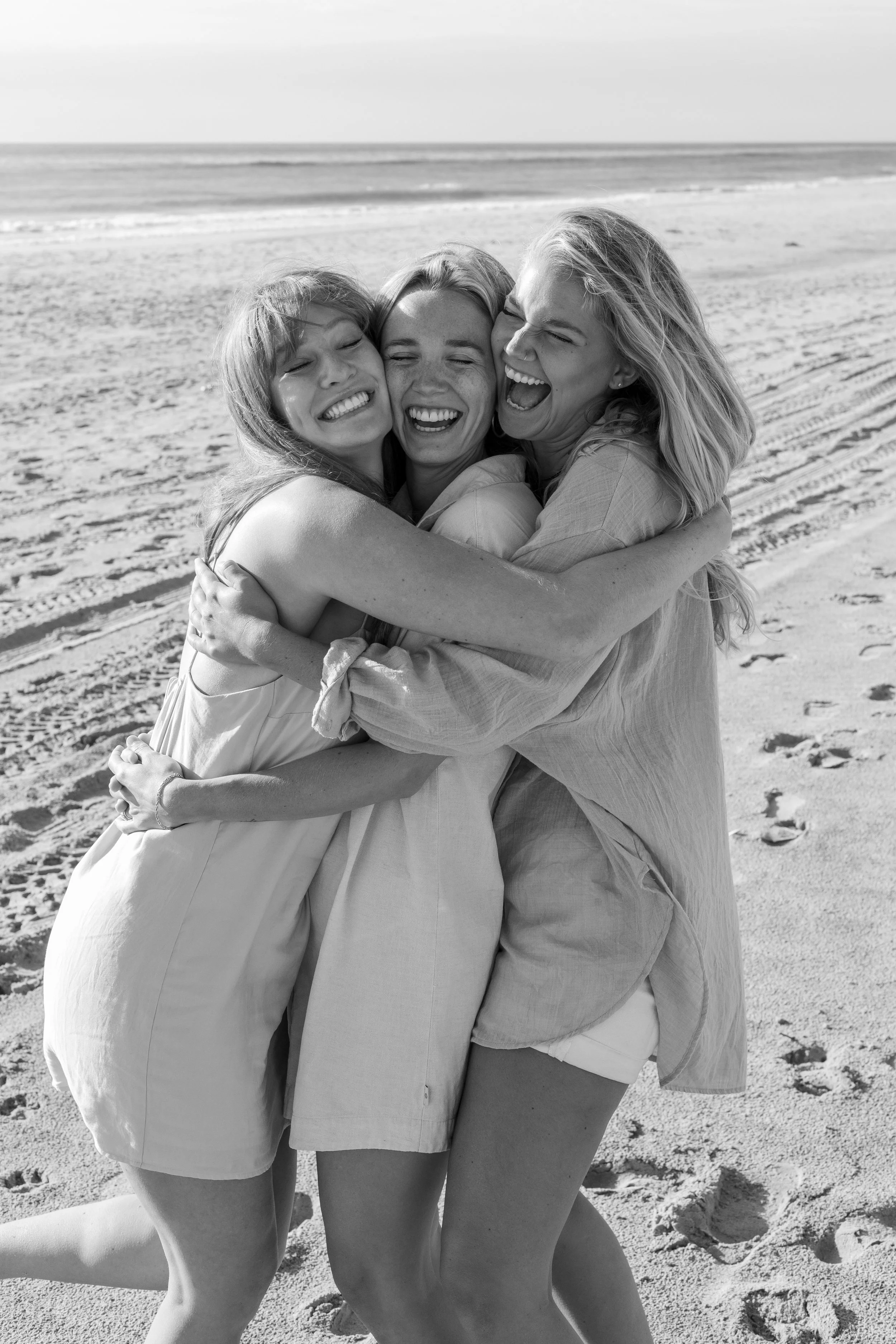 Three women hugging and laughing on a sandy beach, black and white photo.