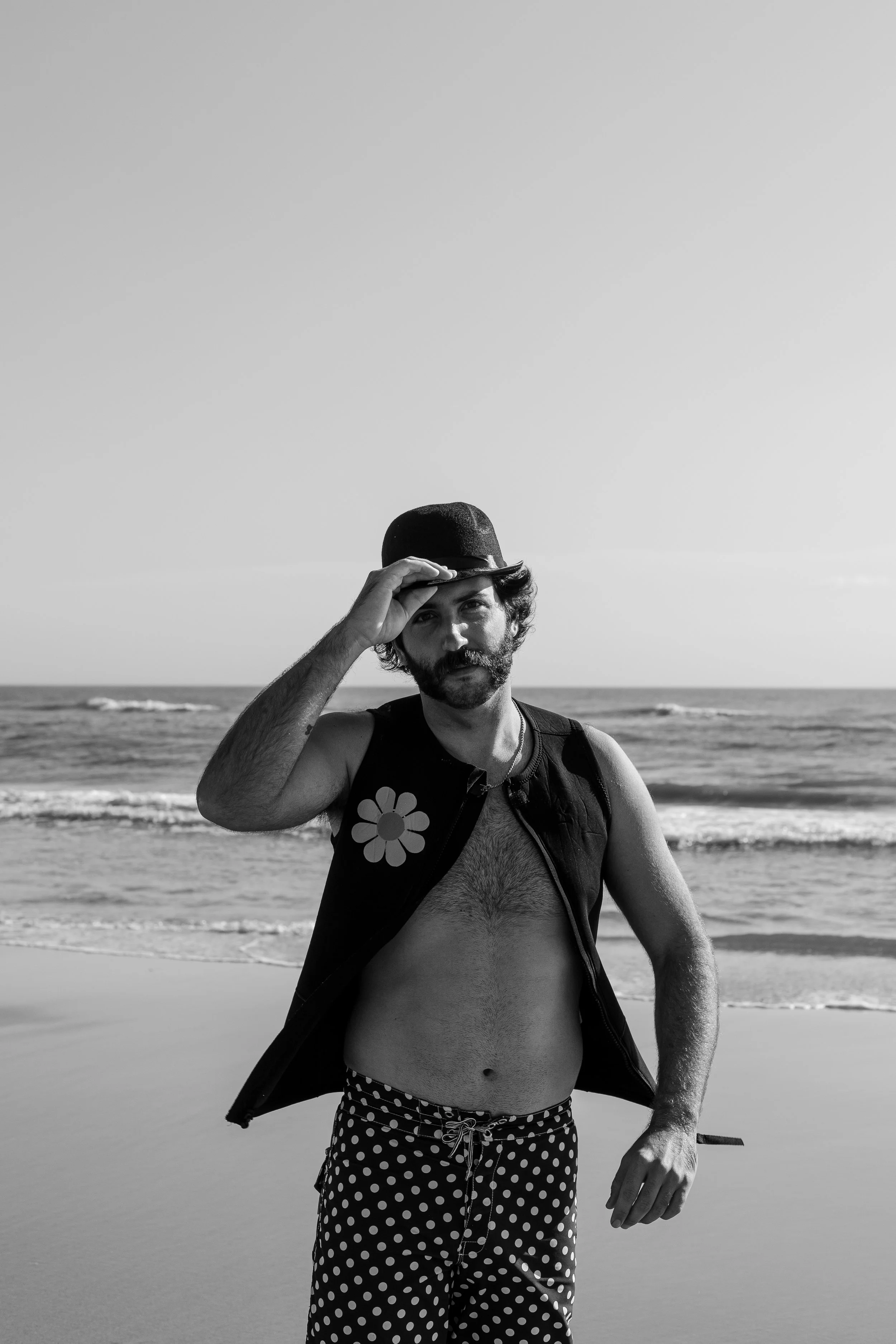 A man with a beard and mustache, wearing polka dot swim trunks, a sleeveless vest with a flower patch, a necklace, and a cap, stands on a beach with ocean waves in the background, tipping his cap and looking at the camera.