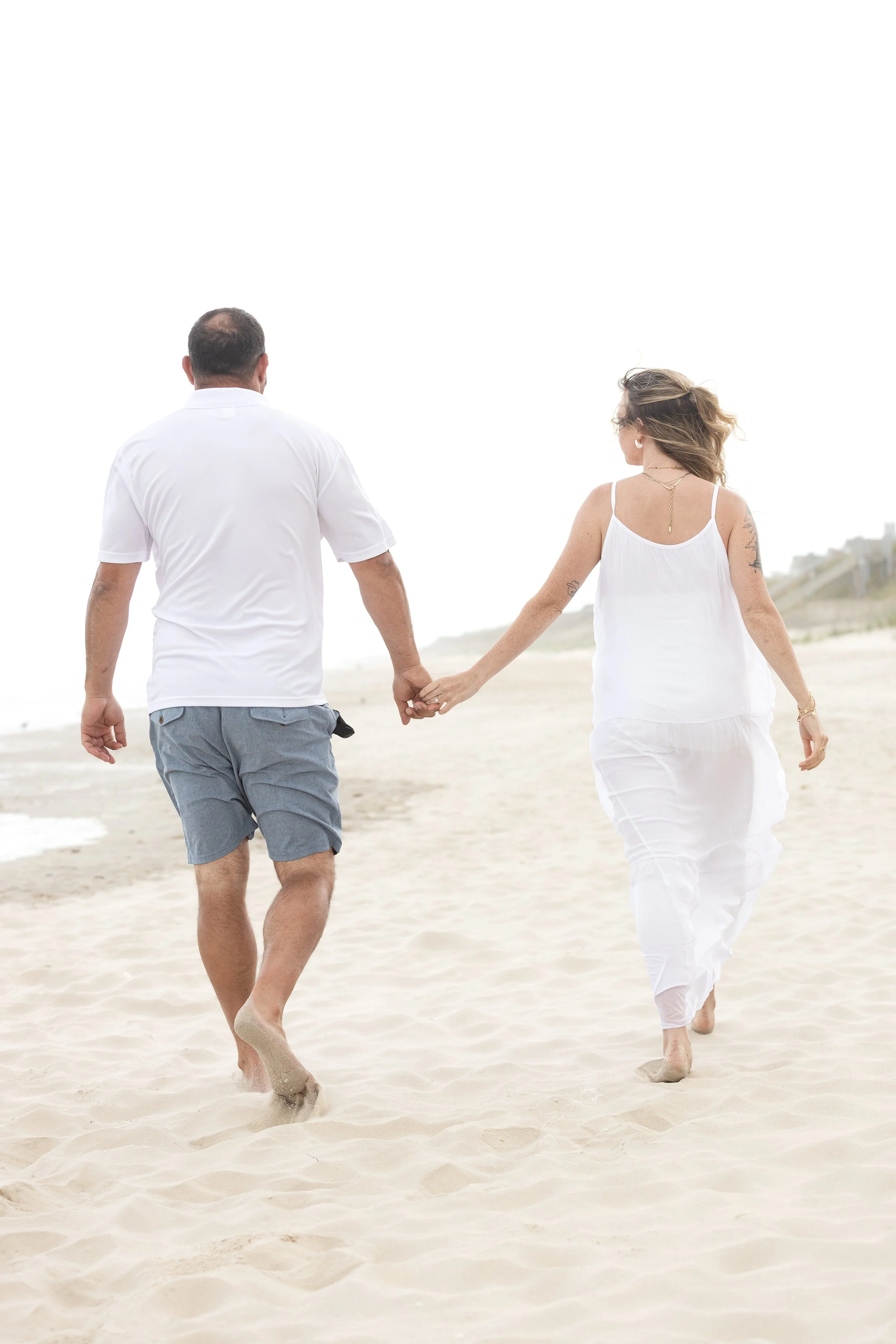 A couple walking hand in hand on a sandy beach, dressed in casual white clothing with the ocean and sky in the background.