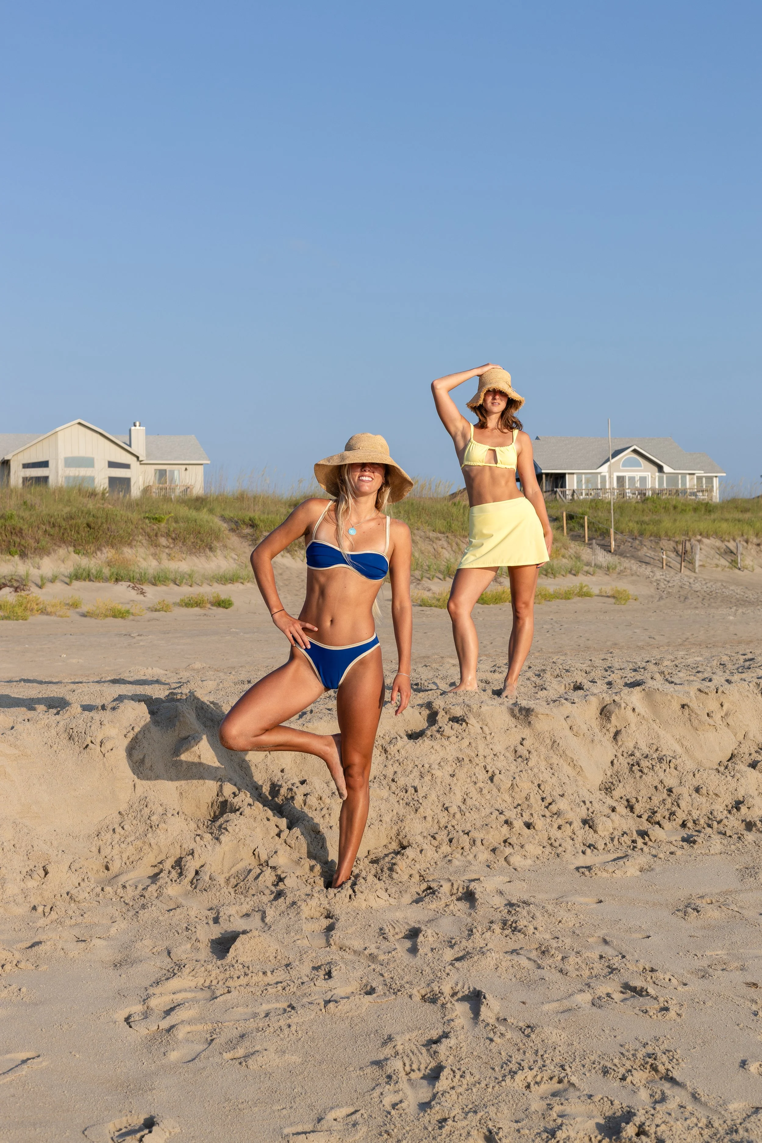 Two women in swimsuits posing on a sandy beach, wearing large straw hats and smiling, with houses and grassy dunes in the background.