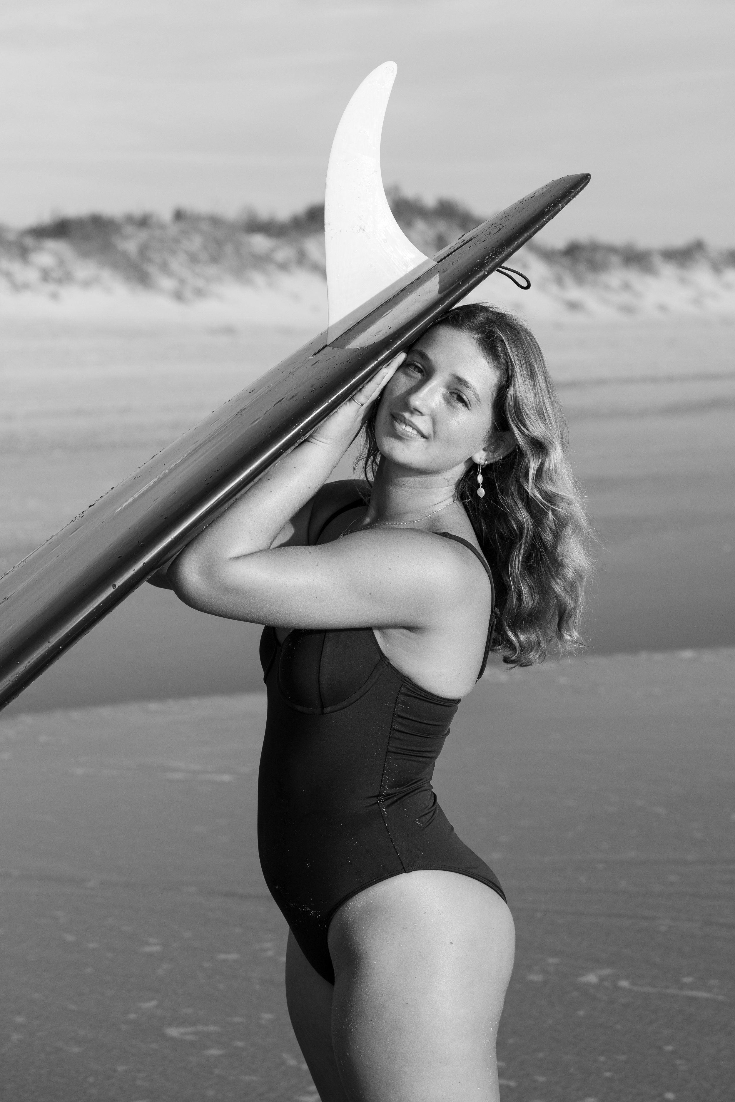 A young woman in a black swimsuit holding a surfboard at the beach, with sand dunes and the ocean in the background.