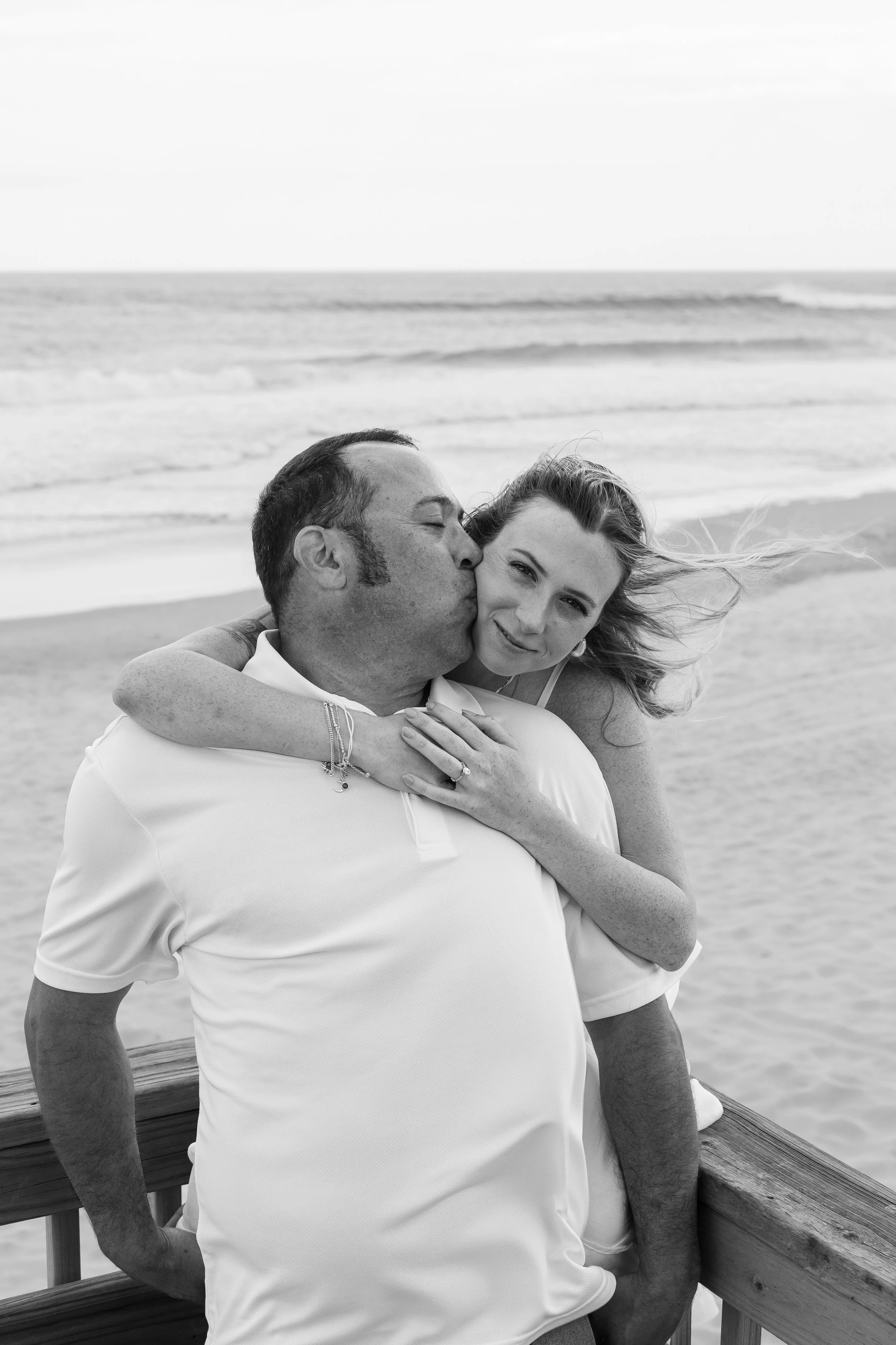 A man kissing a woman on the cheek while she smiles, on a beach with the ocean and waves in the background.