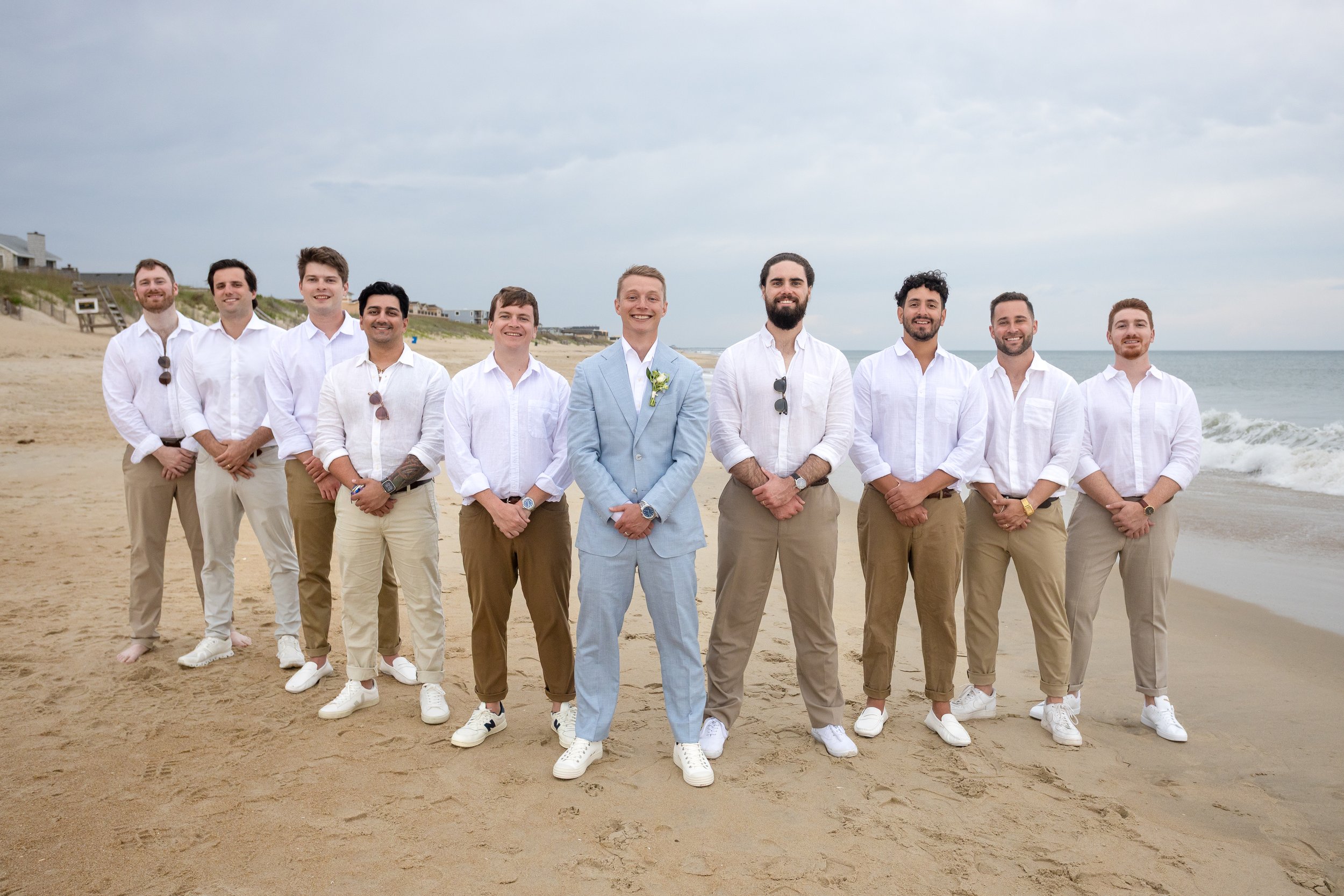 Group of men in white shirts and beige pants standing on a beach with ocean waves and cloudy sky in the background.