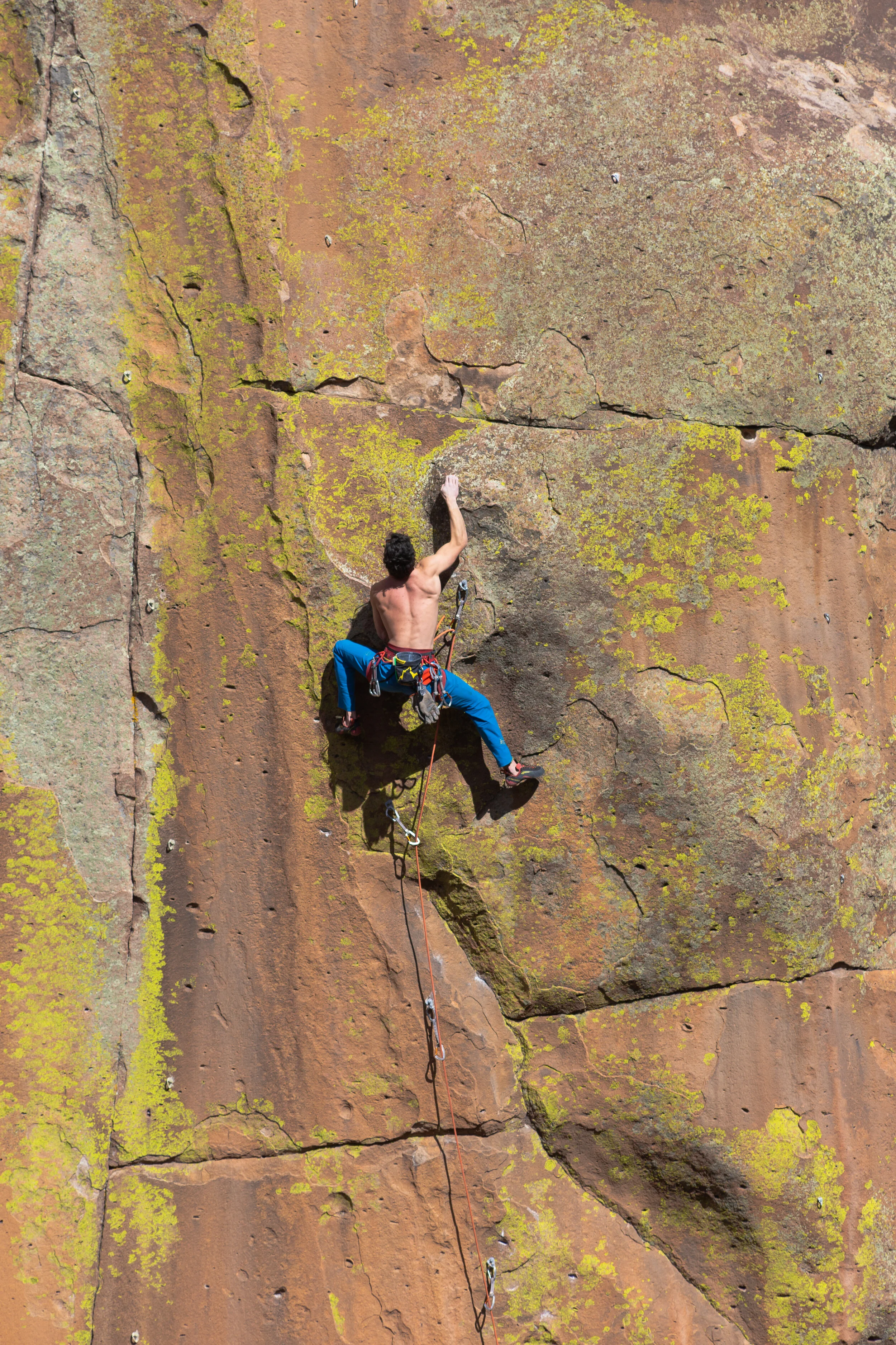 A male rock climber scaling a steep outdoor rock face with moss and cracks, wearing blue climbing pants and harness.