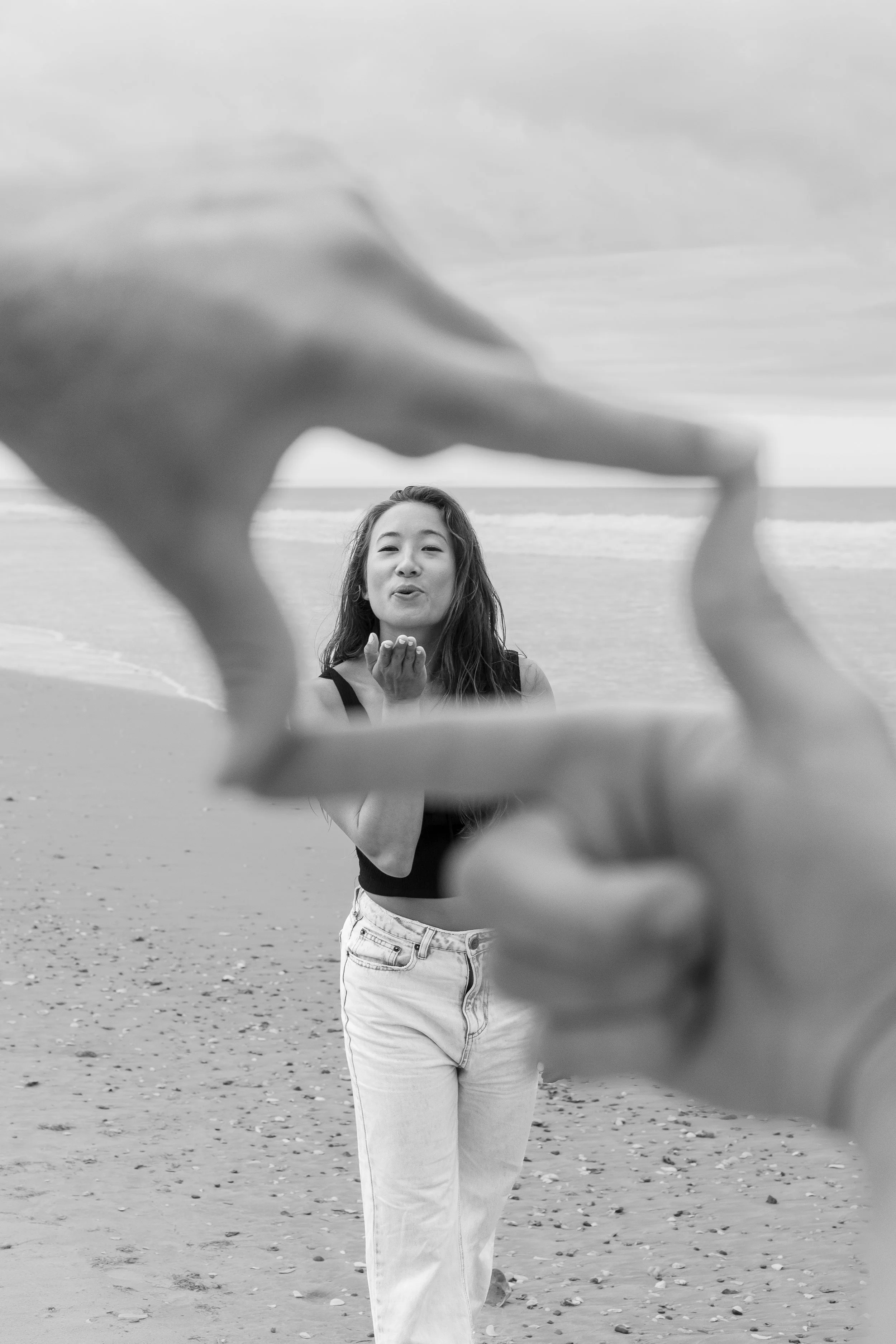 A woman on a beach blowing a kiss, seen through a person's hand frame in the foreground.