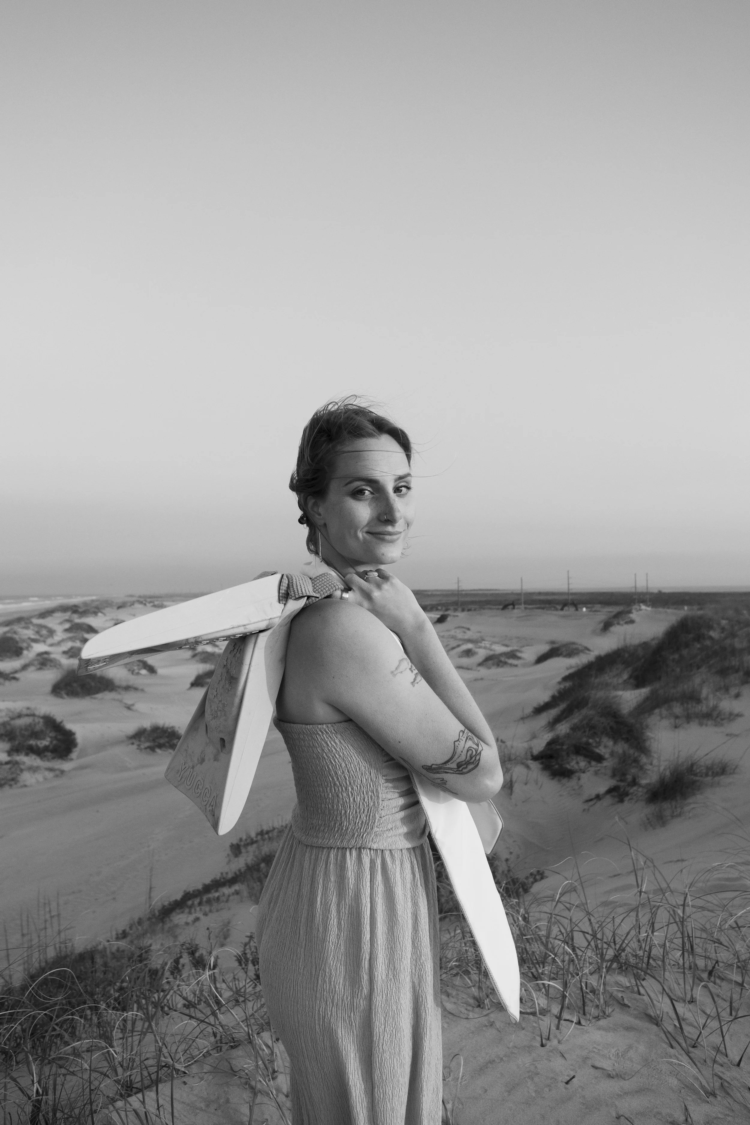 A woman with tattoos standing on sandy dunes at the beach, holding a skateboard over her shoulder, in a black-and-white photo.
