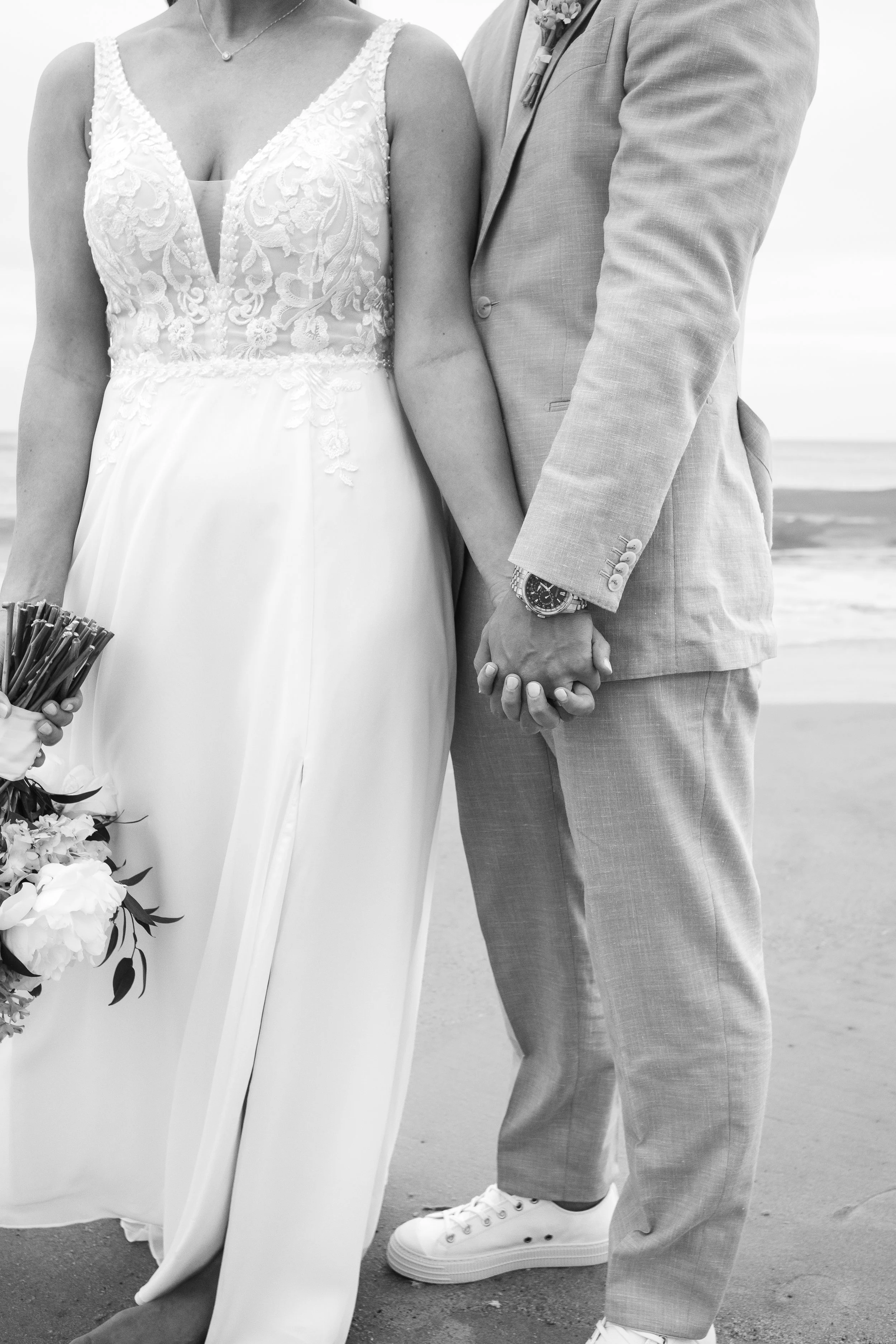 A bride and groom holding hands on the beach, close-up of their torsos and hands, with the bride wearing a lace wedding dress and the groom in a light suit, holding hands with wedding rings visible.