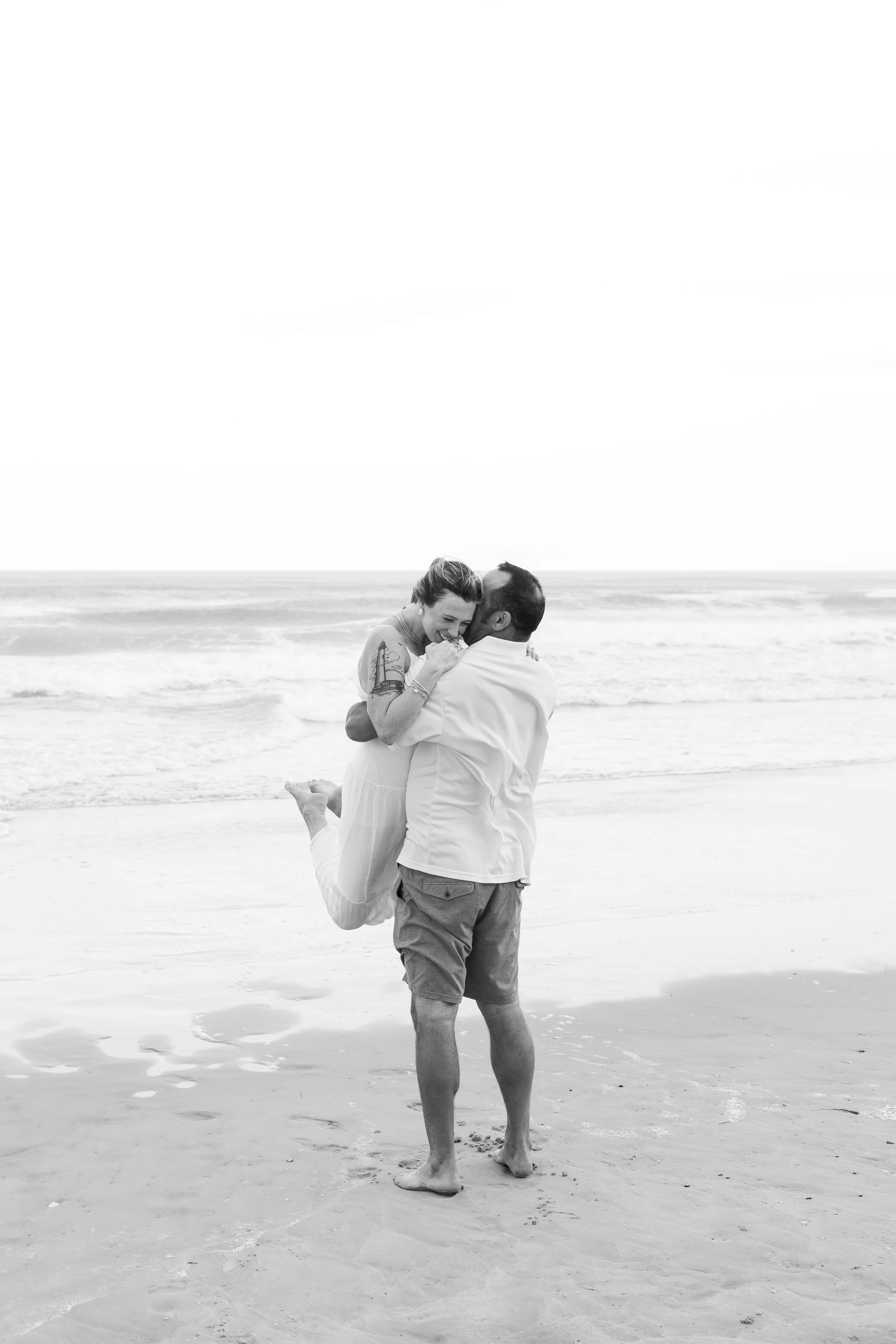 A couple on the beach, with the man lifting the woman in his arms, both smiling and enjoying a romantic moment.