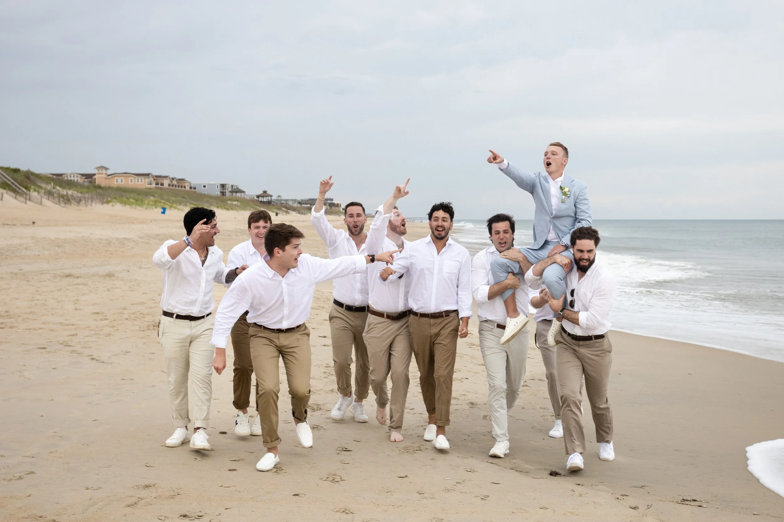 A group of men wearing white shirts and beige pants on a beach, with one man in a light blue suit sitting on another man's shoulders, all celebrating and pointing towards the sky.