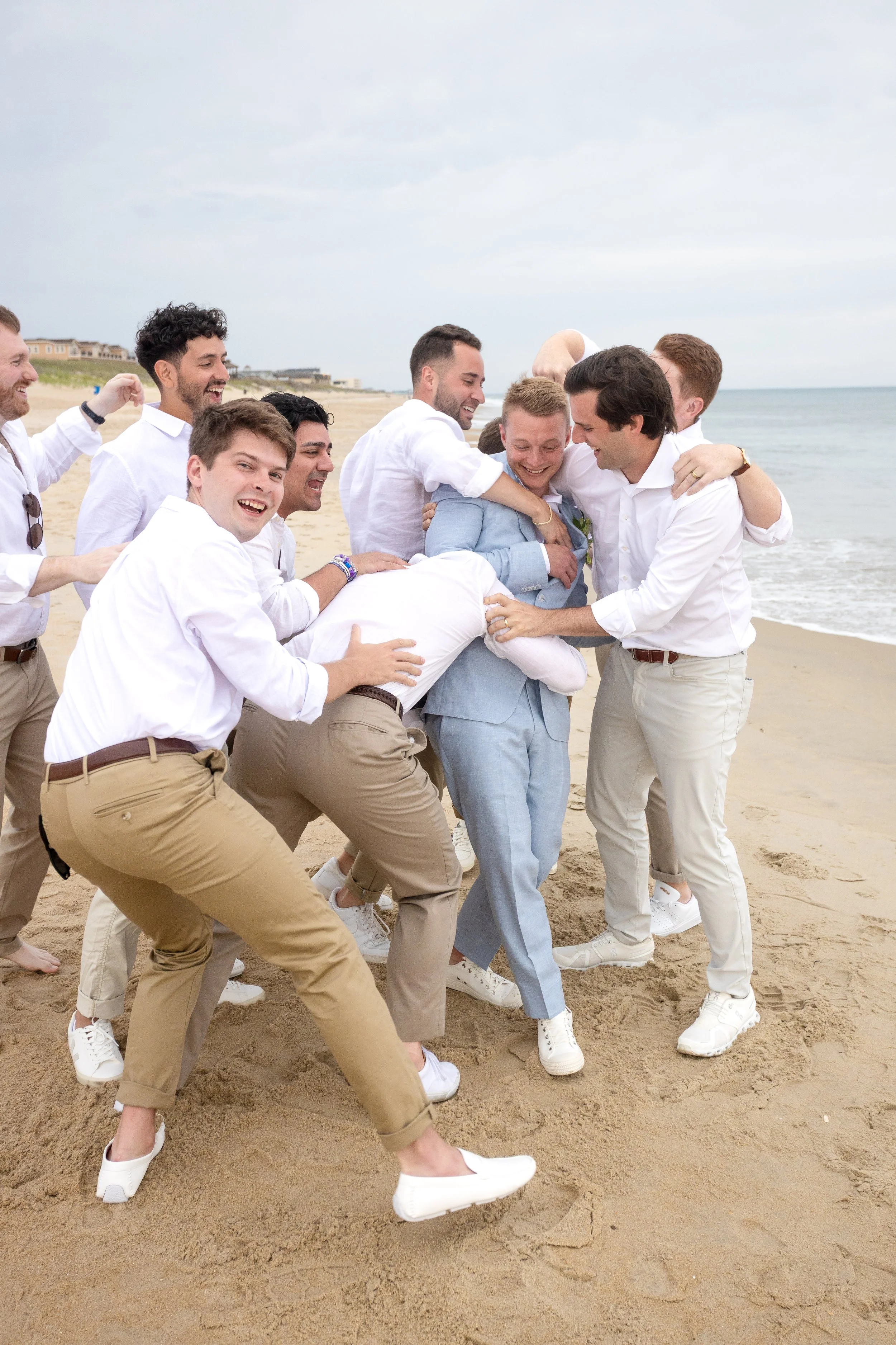 Group of men wearing white shirts and khakis or beige pants playfully tugging a man in a light blue suit on the beach.