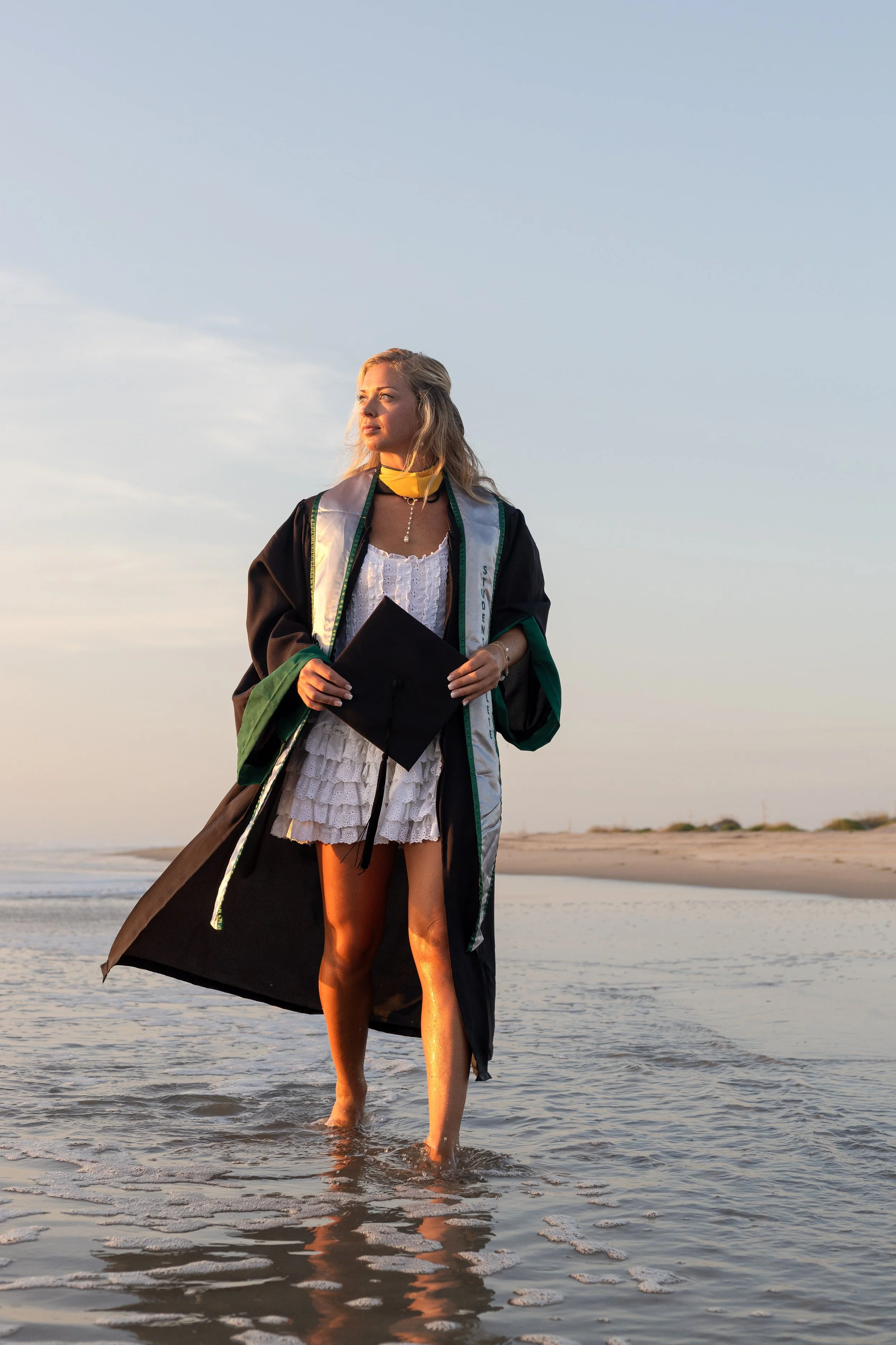 A woman in a graduation gown holding a diploma stands barefoot in shallow ocean water on the beach during sunset.