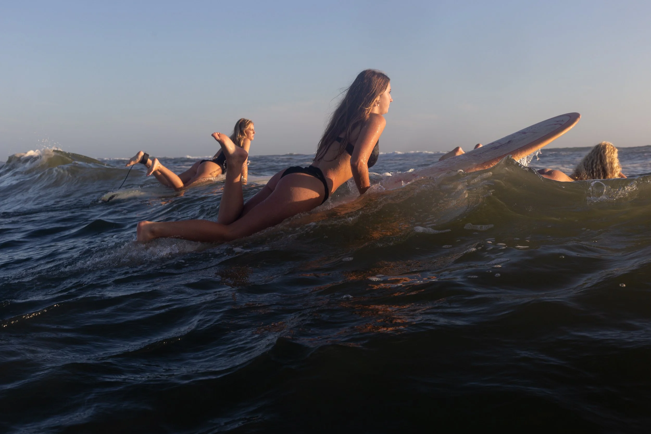 Three women surfing in the ocean, one woman in the foreground wearing a black bikini, on a surfboard, paddling and looking ahead, during sunset or sunrise.