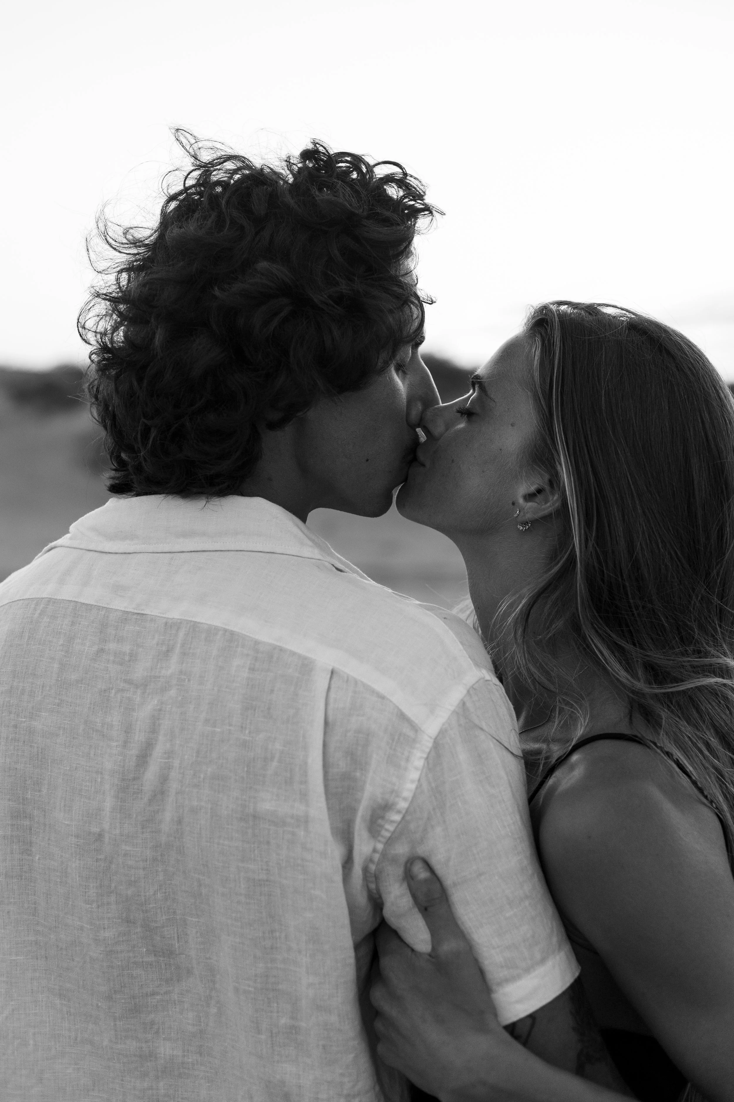 A black-and-white photo of a couple kissing outdoors, with the woman holding the man's shoulder, both with their eyes closed.