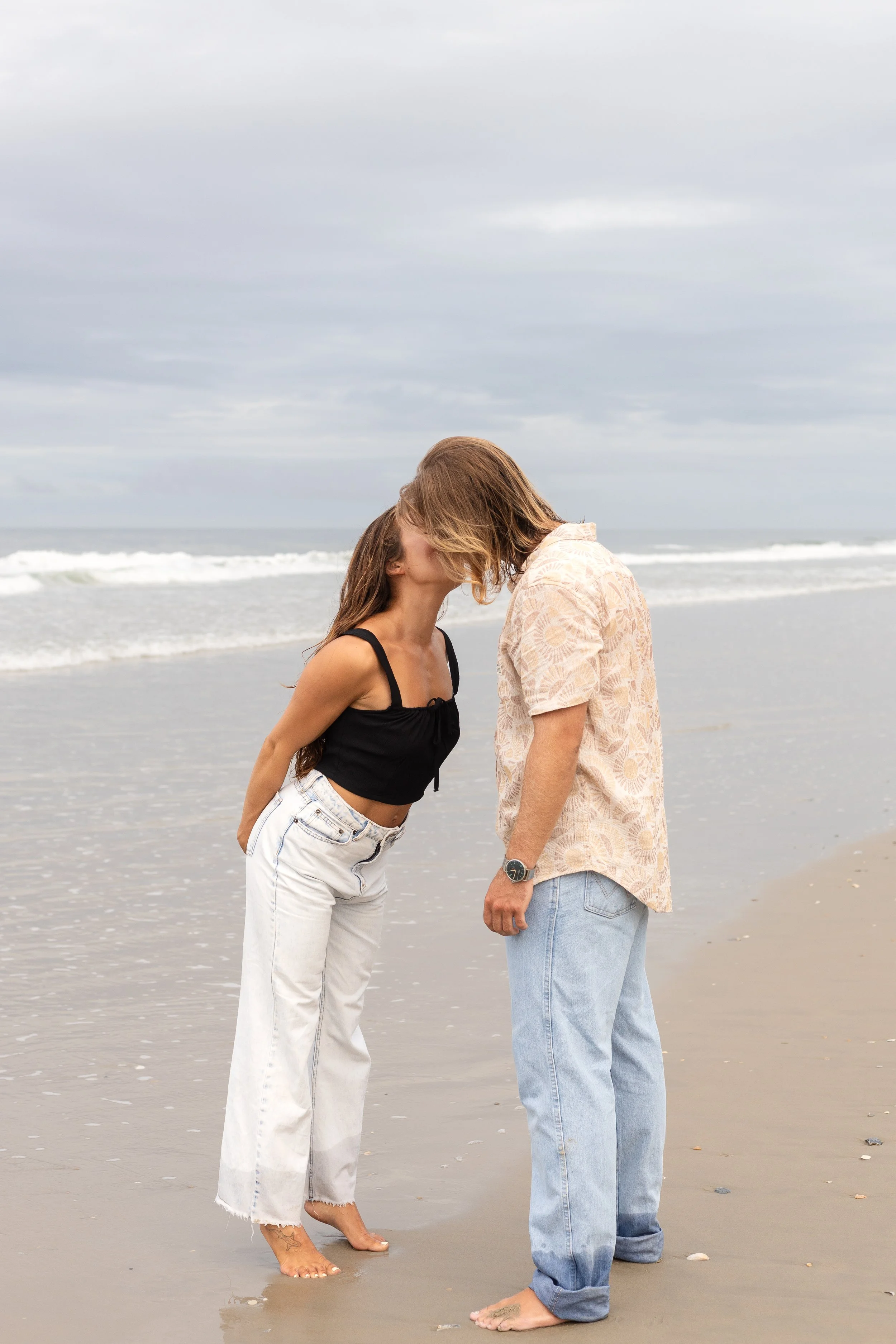 A young woman and a young man kissing on the beach, standing barefoot near the shoreline with a cloudy sky overhead.