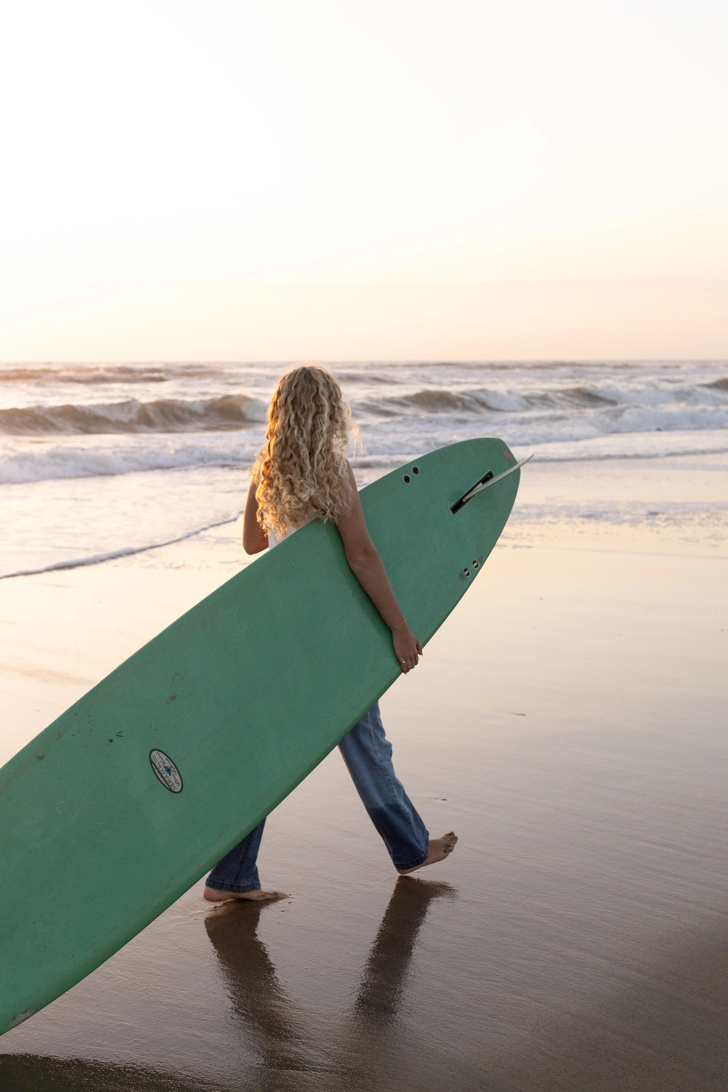 A woman with curly blonde hair carrying a surfboard while walking on the wet sand at the beach during sunset.