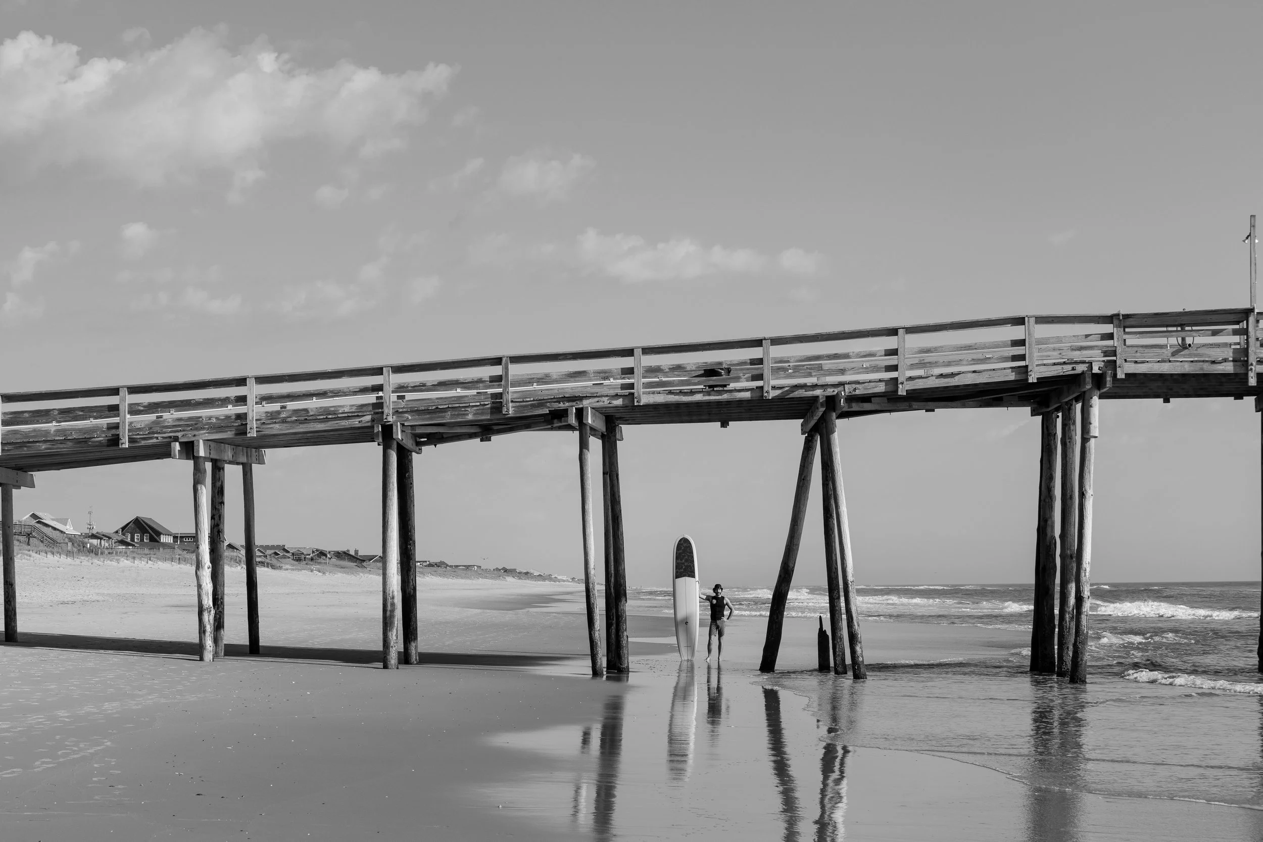Black and white photo of a person standing with a surfboard under a wooden pier on a beach, with ocean waves in the background.