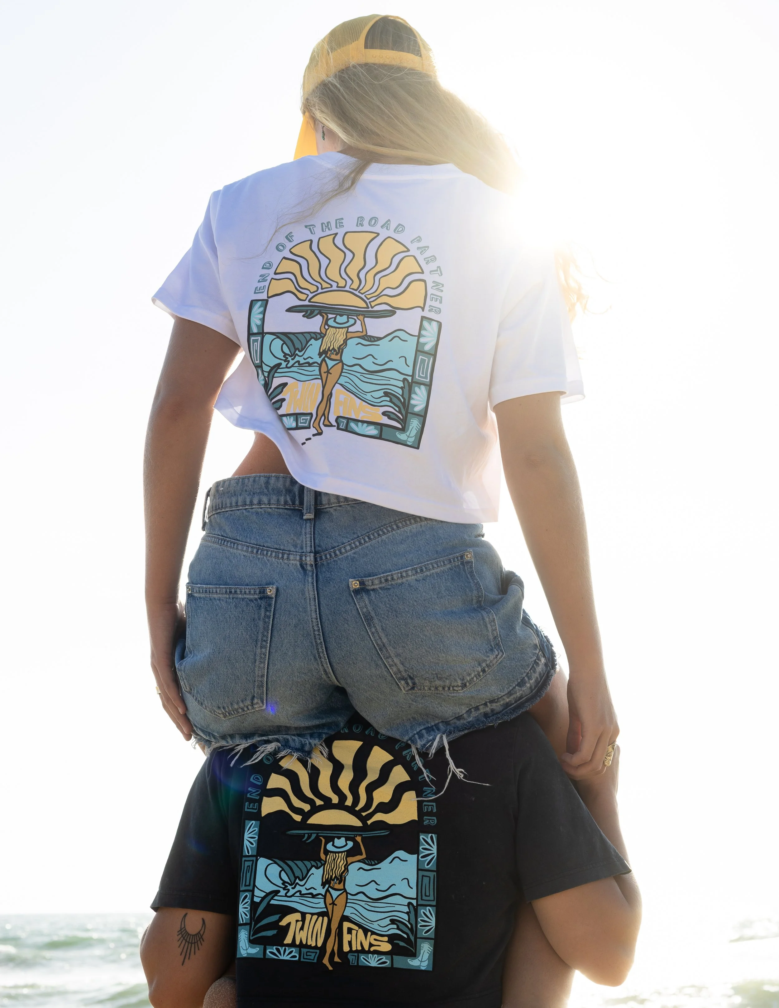 A person standing on the shoulders of another at the beach, both wearing matching T-shirts with a sun and ocean graphic and the words "End of the Road" and "Two Fins."