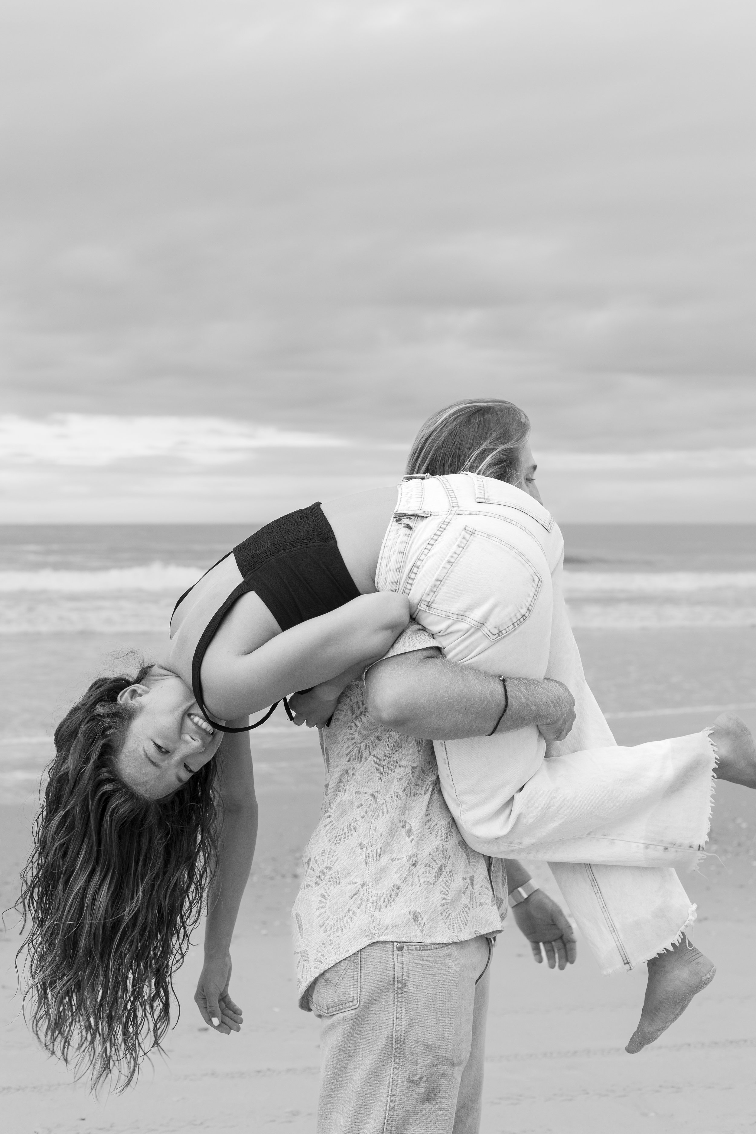A woman is giving a piggyback ride to a girl at the beach. The girl is hanging upside down, smiling, and the scene is overcast.