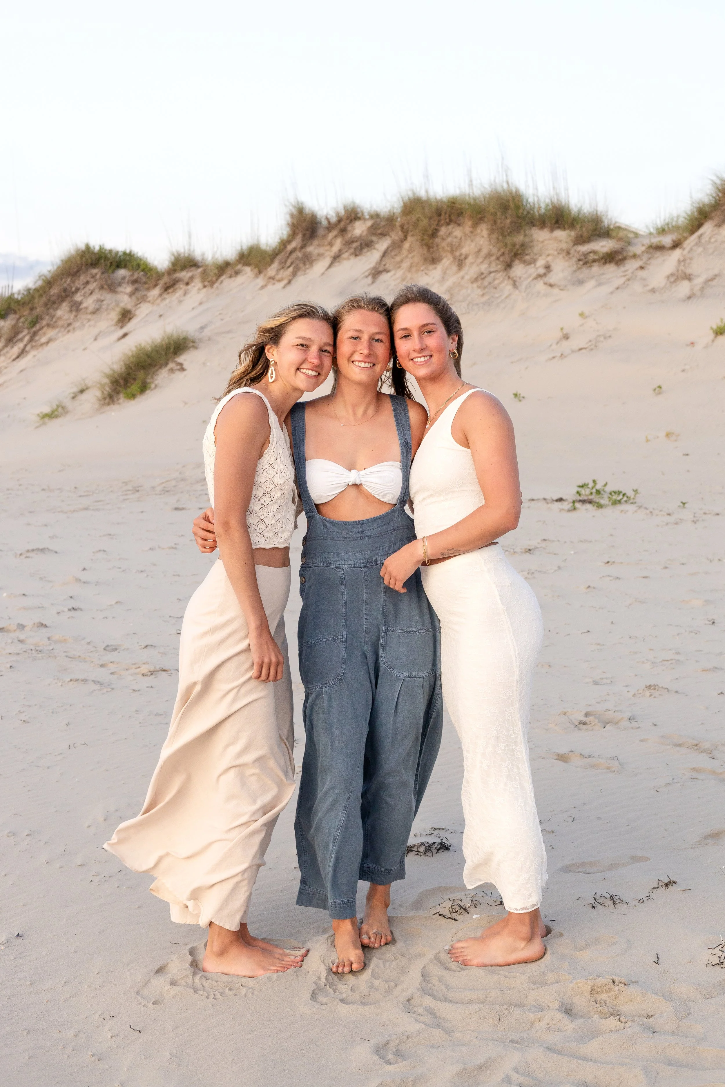 Three women standing on a sandy beach near dunes, smiling at the camera.