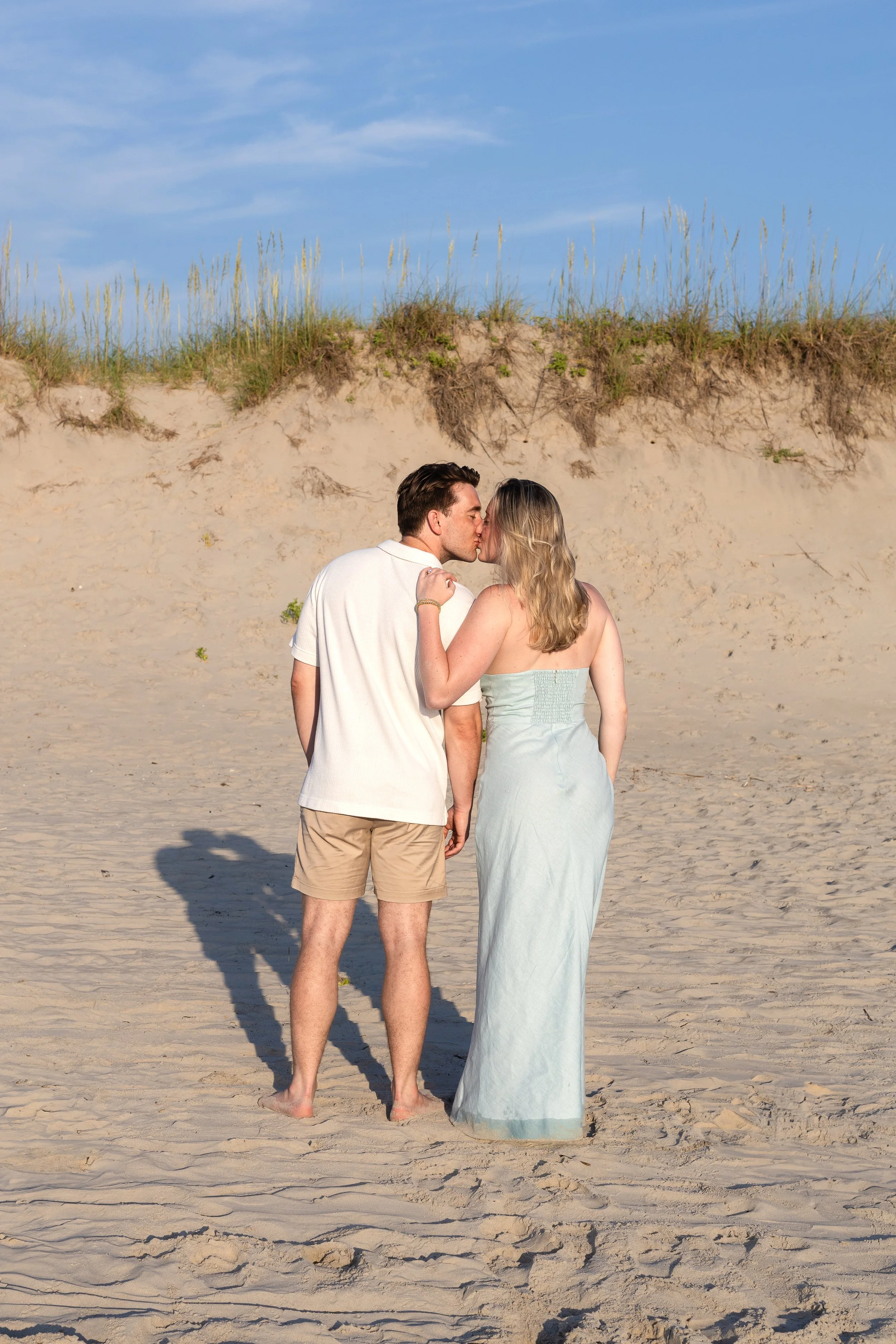 A couple kissing on the beach, standing barefoot on sand with a background of sand dunes and blue sky.