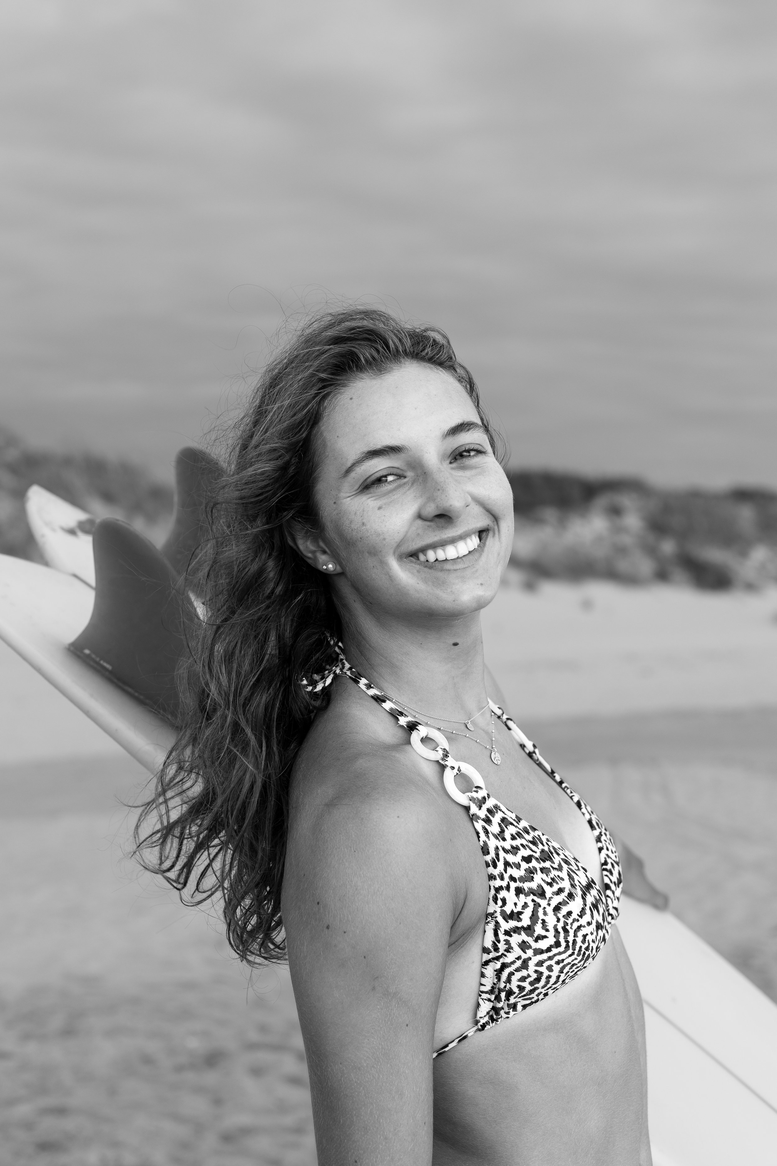 A young woman smiling on a beach, holding a surfboard, wearing a patterned bikini top.