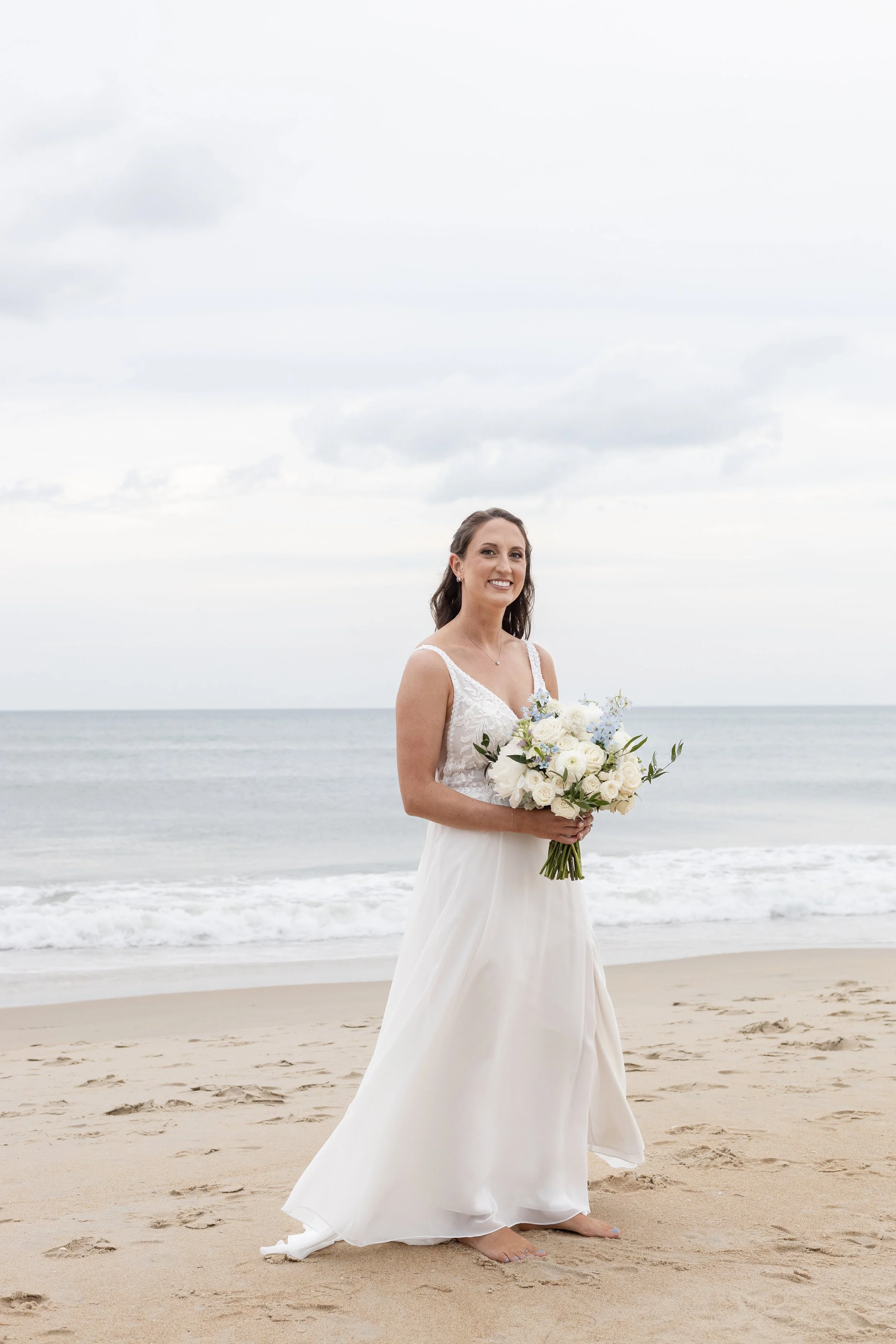 A bride in a white wedding dress holding a bouquet of white and light blue flowers, standing barefoot on the sandy beach with the ocean in the background and a cloudy sky overhead.