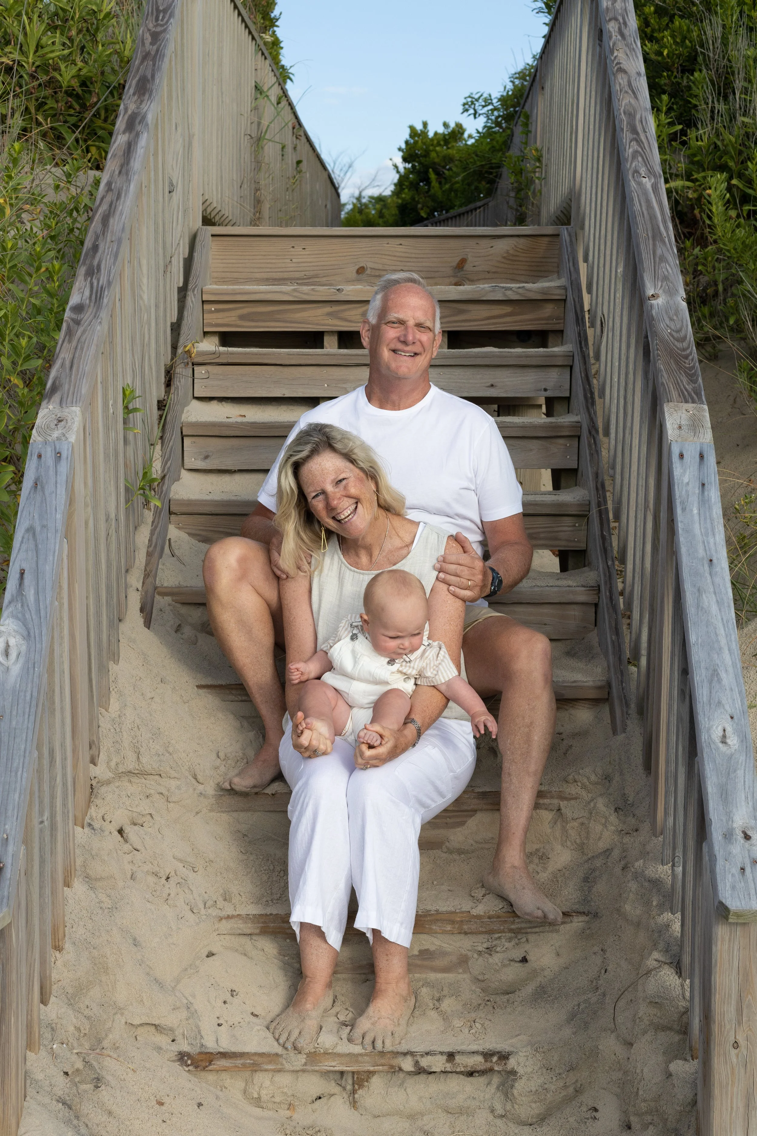 A happy family of three sitting on beach stairs with sand at their feet. The older woman is sitting on the stairs, holding a baby, with a man sitting behind her. They are outdoors on a path with greenery on either side.