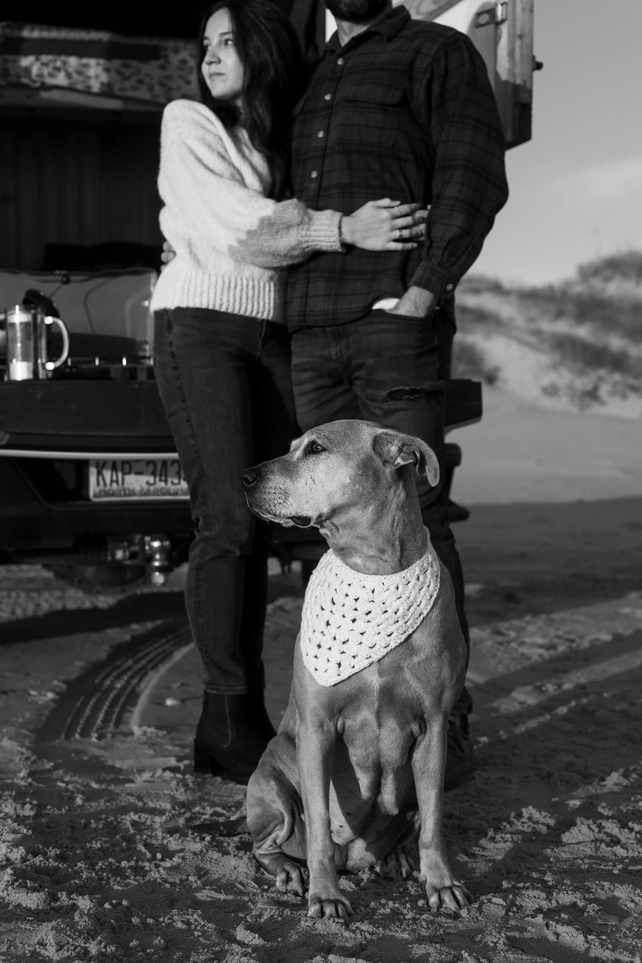 A black and white photo of a couple with their dog on a sandy beach. The dog, a large breed, is sitting in the sand wearing a knitted bandana. The woman is leaning against the man, who is standing in front of a car with a trailer, with a background o