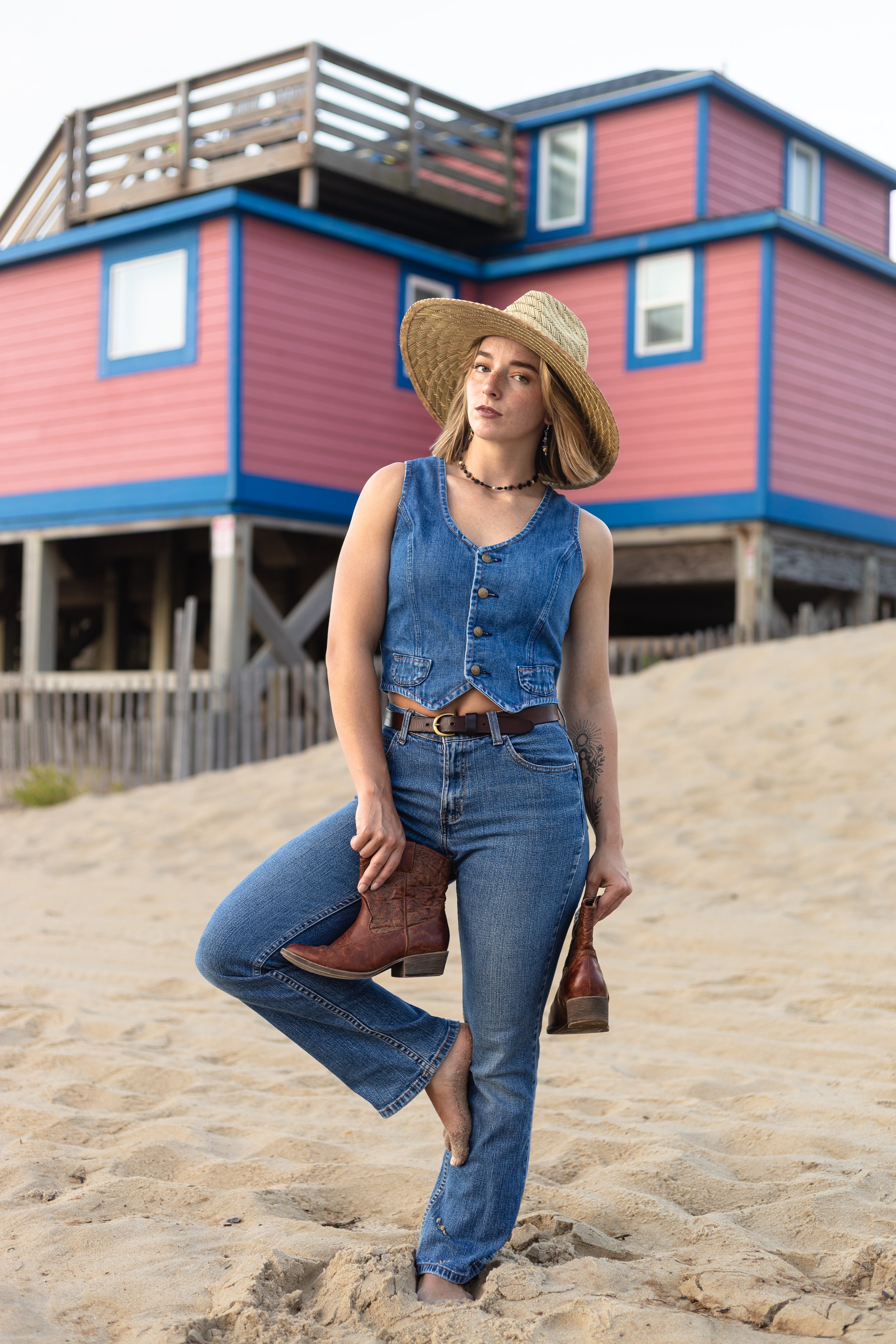 A young woman in denim outfit and wide-brimmed hat standing on sandy beach near colorful house, holding cowboy boots.