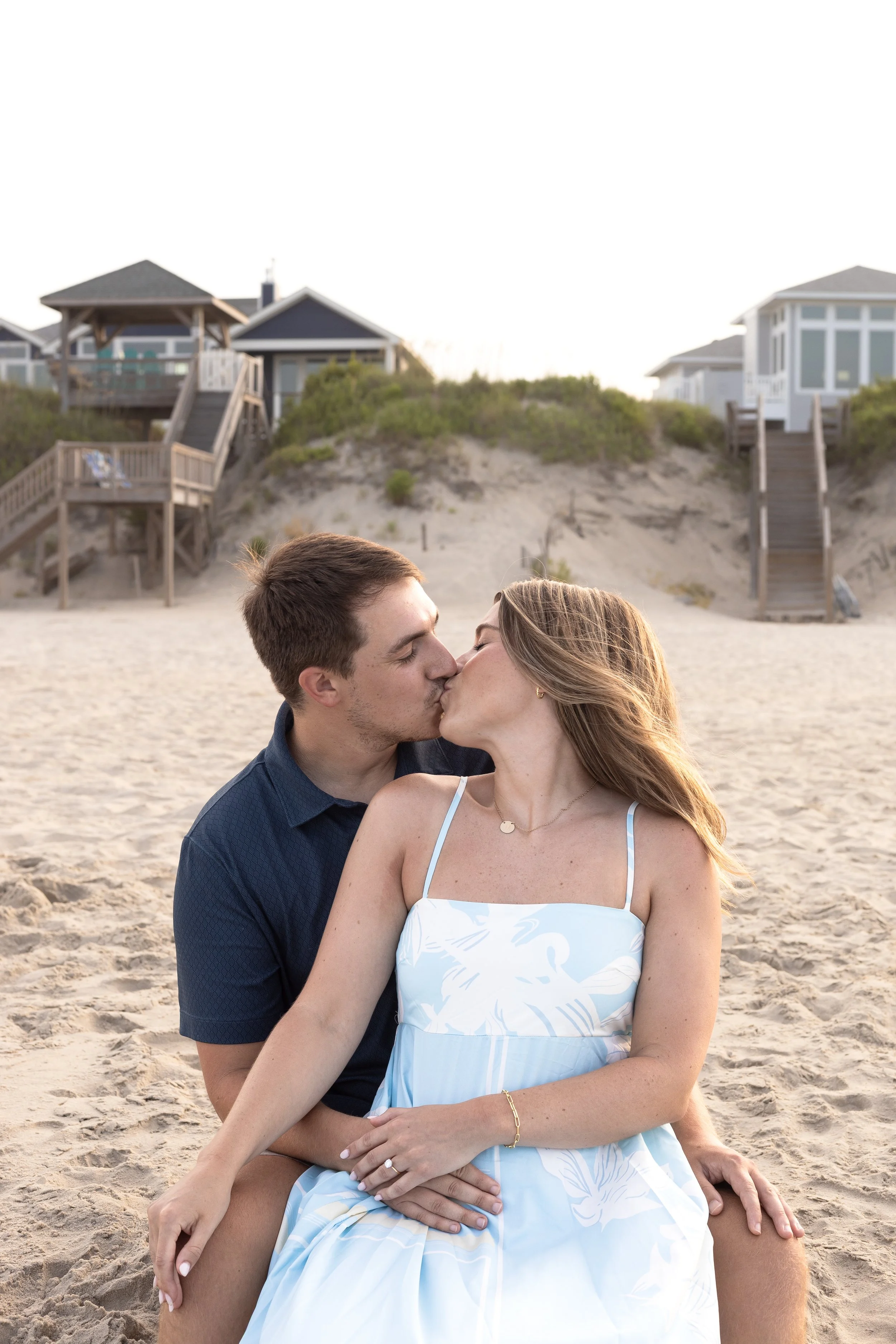 A couple kissing on the beach, with houses and stairs on dunes in the background.