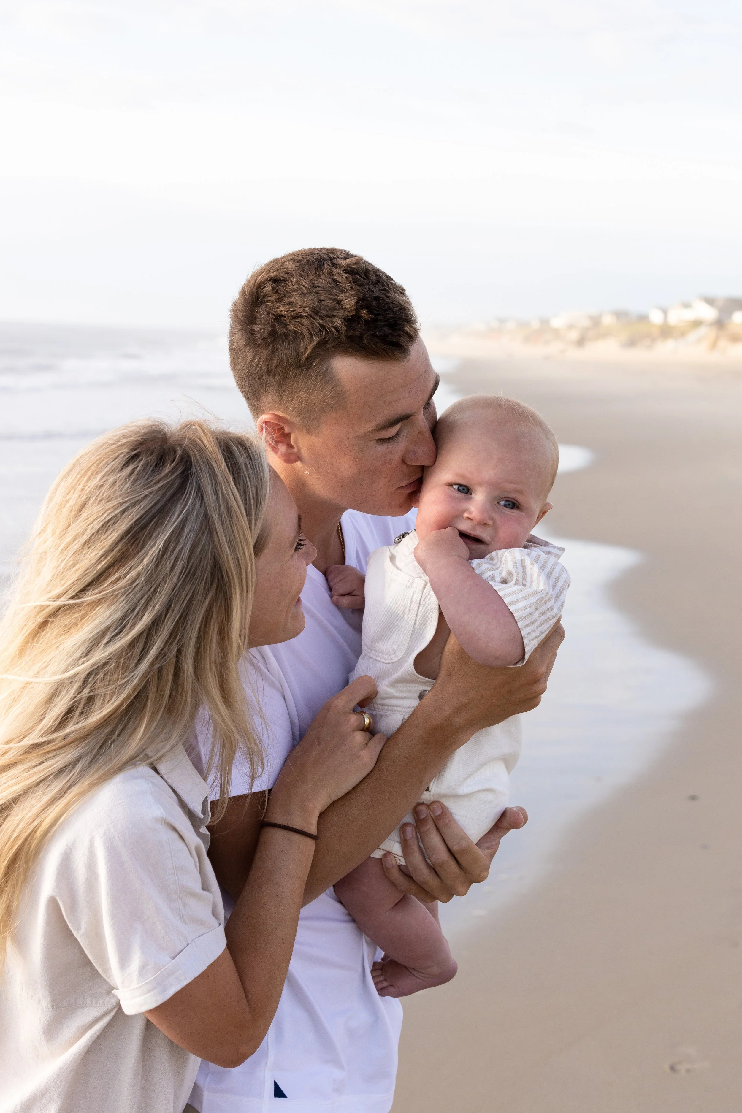 A young family of three on the beach, with the father holding the baby and the mother gently touching the baby. The father kisses the baby's cheek while the baby looks slightly distressed. The background shows the shoreline and the ocean under a clou