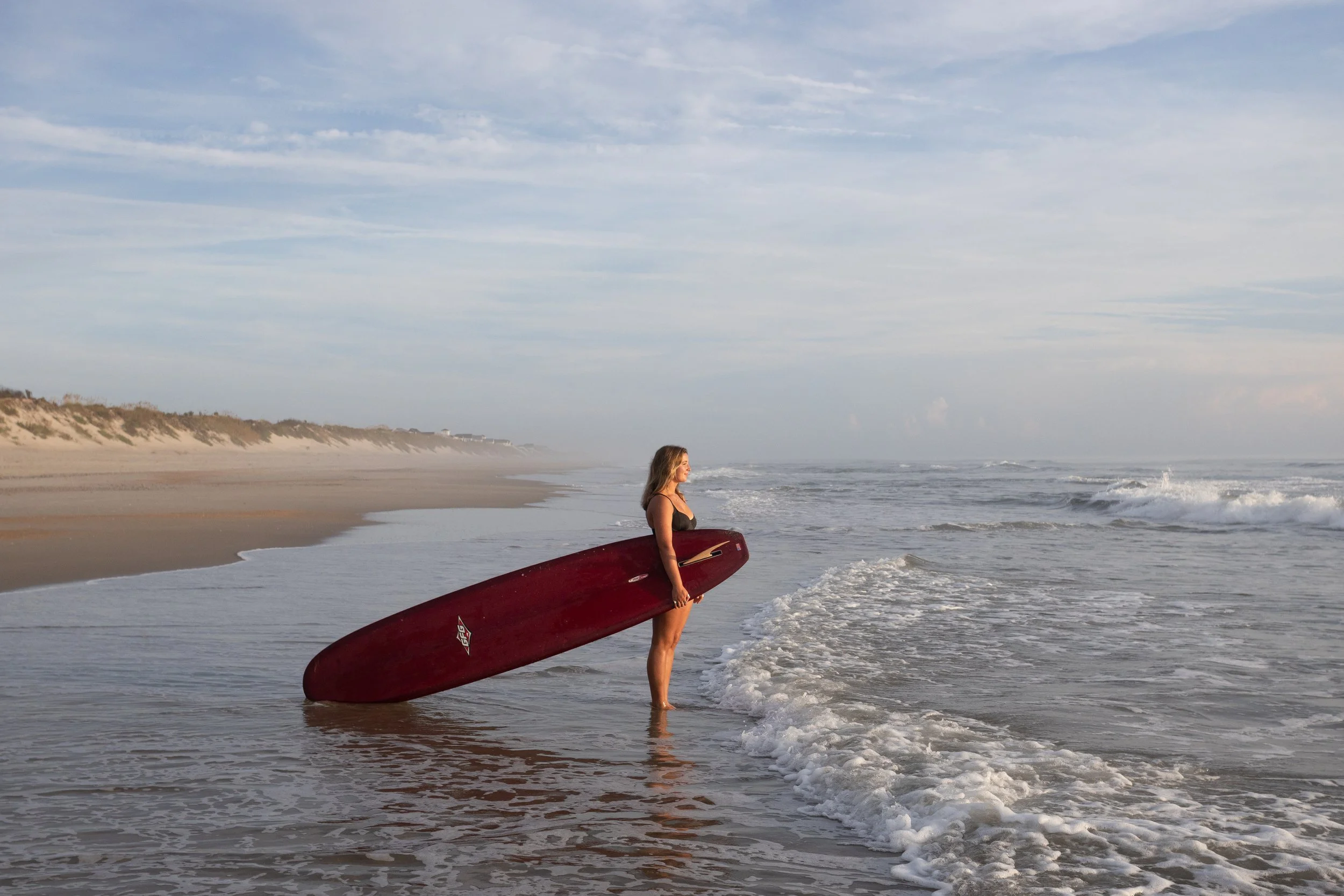 A woman standing ankle-deep in the ocean holding a red surfboard at the beach.