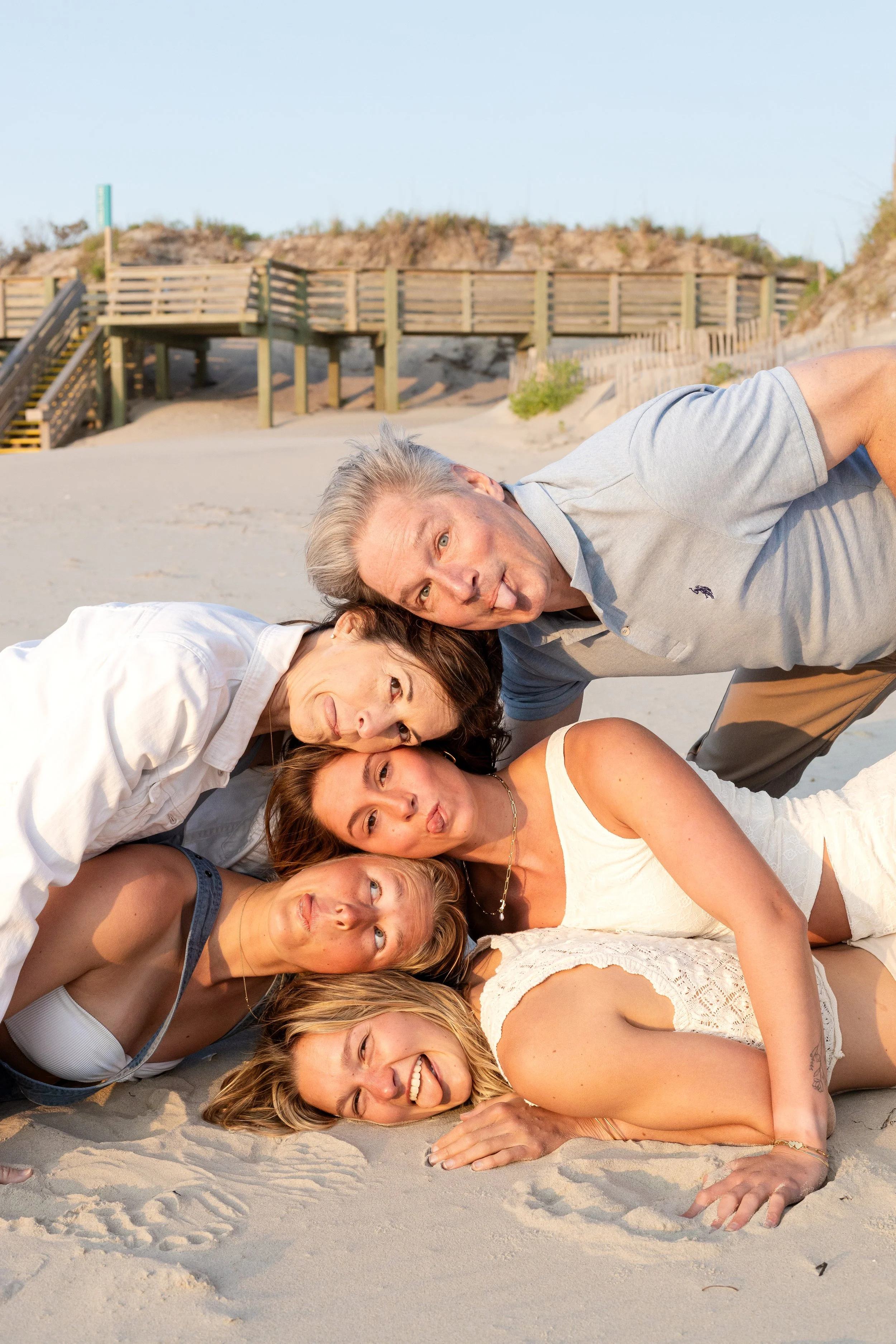 Group of five friends lying on sand at the beach, making funny faces and posing together.