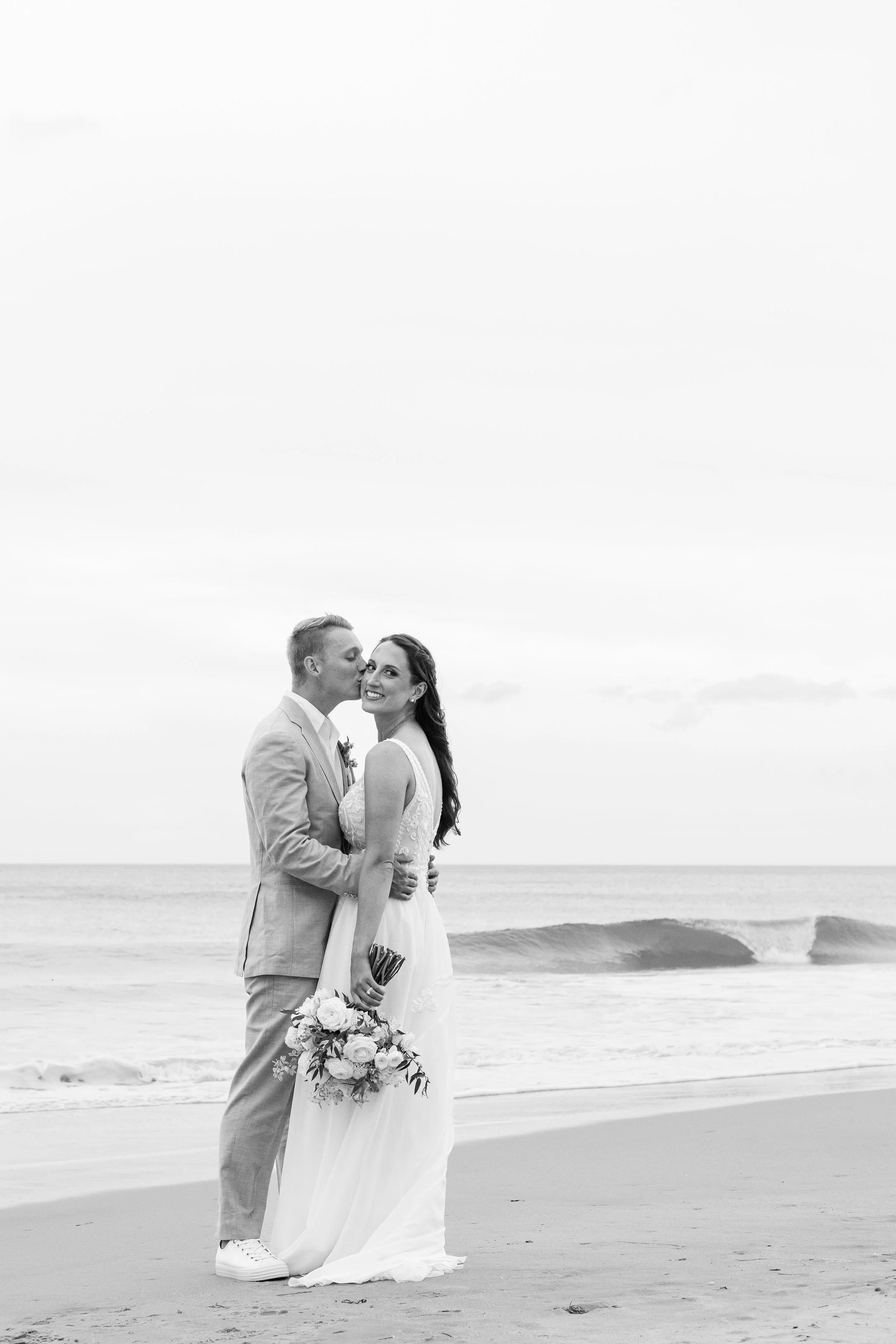 A newlywed couple at the beach, with the groom kissing the bride on the cheek, both smiling. The bride is holding a bouquet of flowers. The scene is in black and white with ocean waves in the background.