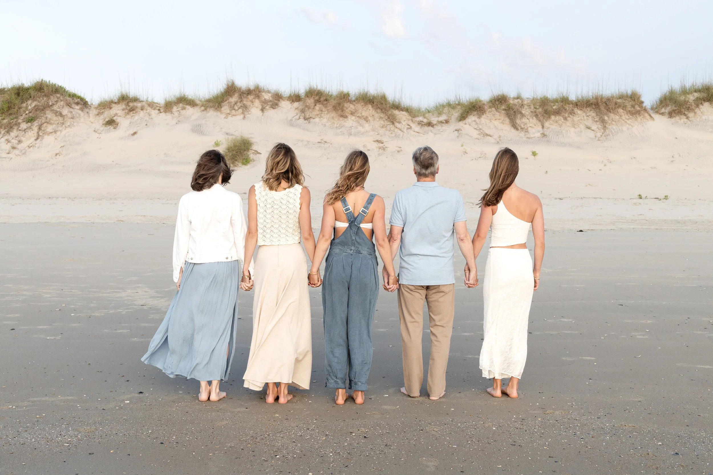 Group of five people holding hands on a beach with sand dunes in the background.