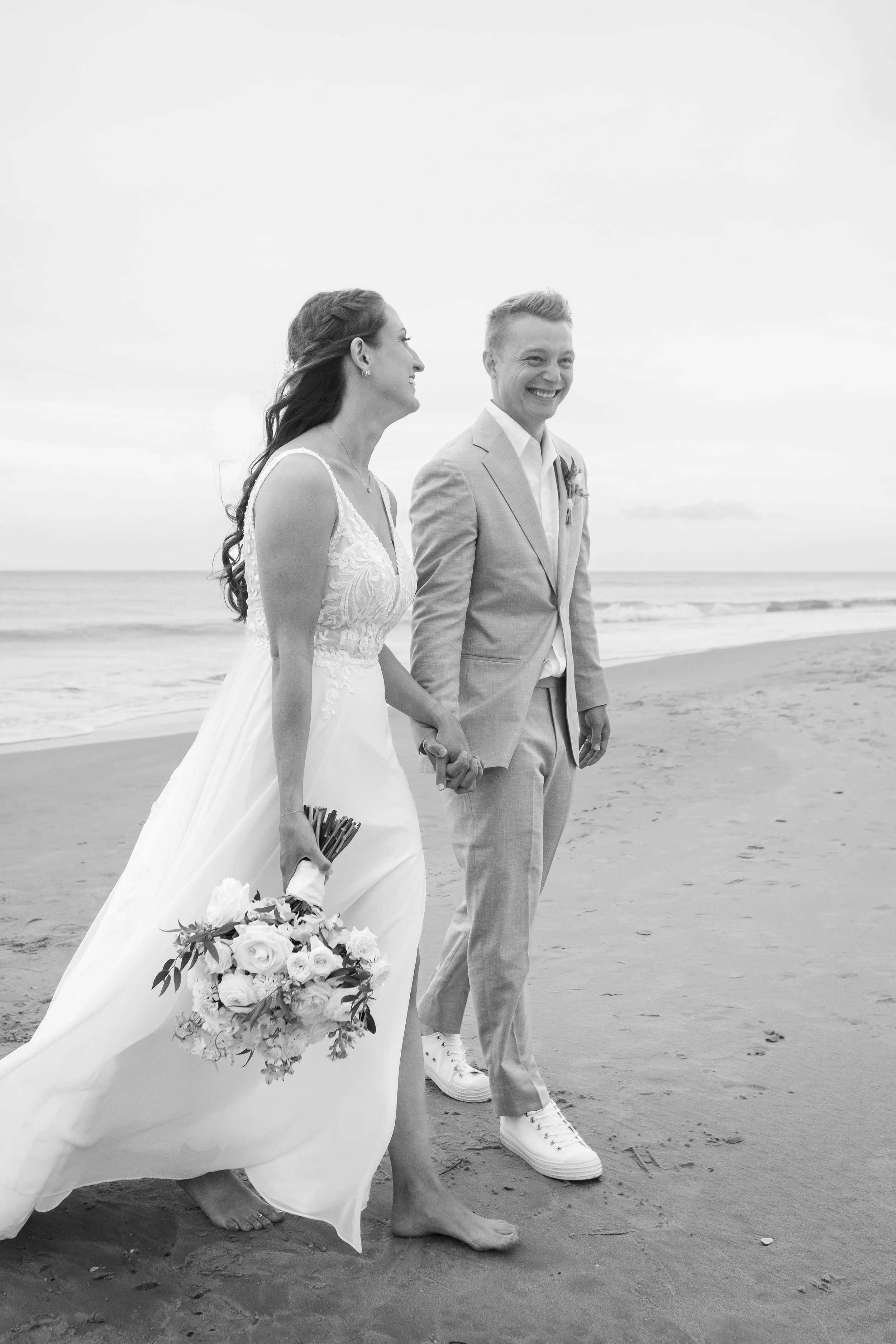 A black and white photo of a bride and groom holding hands on the beach, smiling and walking barefoot.