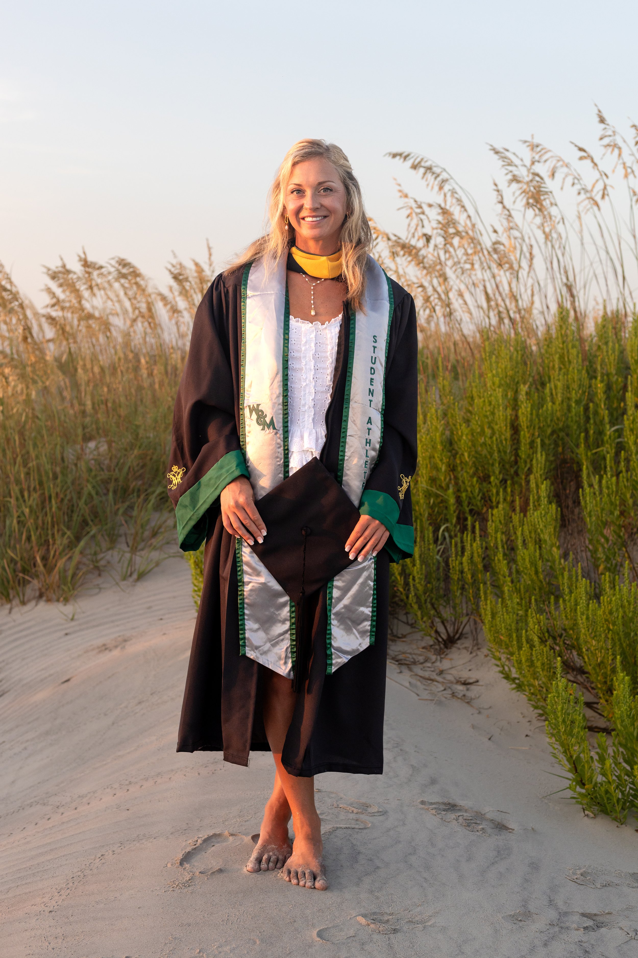 Woman in graduation gown and cap standing barefoot on sandy beach at sunset, holding her cap, with tall grasses in the background.