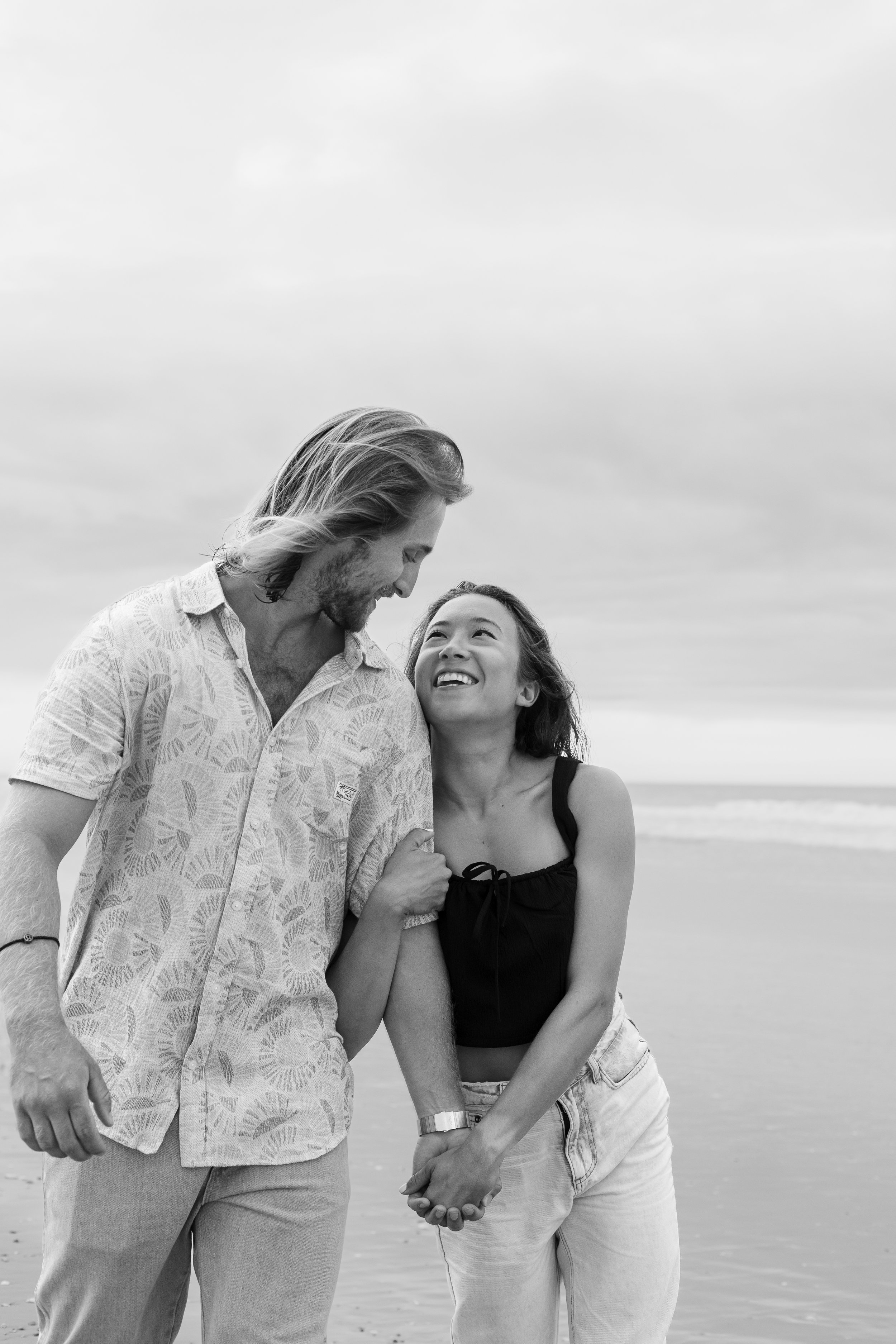 A happy couple holding hands and looking at each other on a beach, smiling, in black and white.