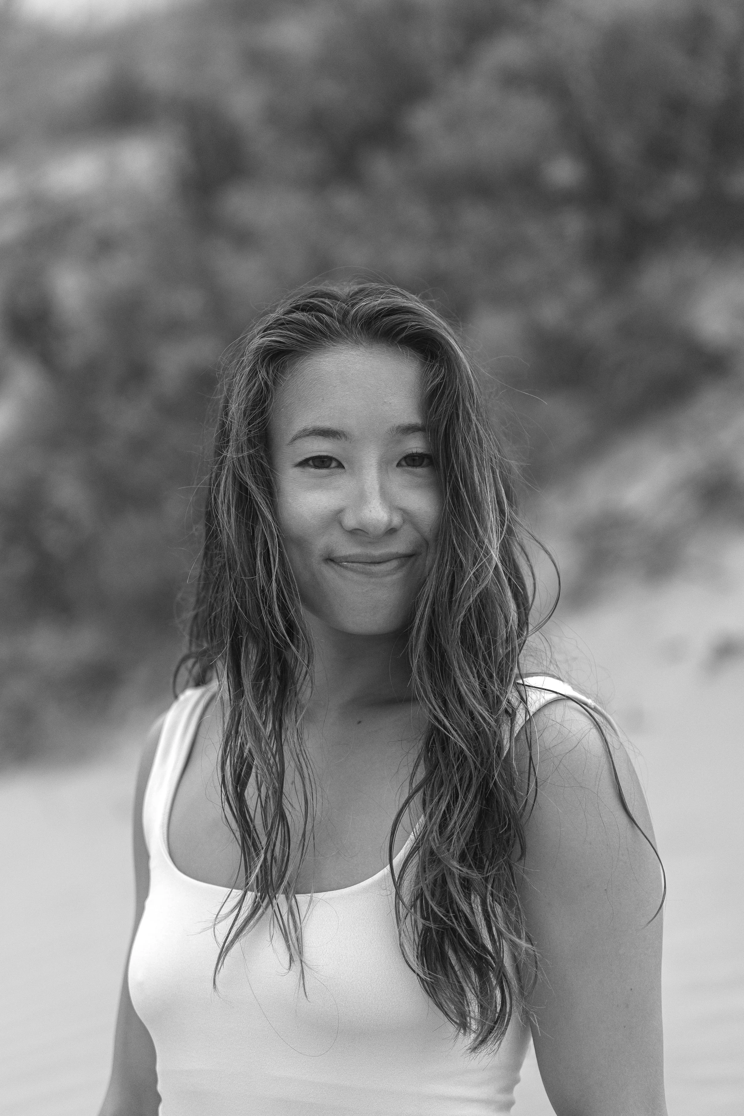 A smiling woman with long, wet hair wearing a white tank top outdoors.