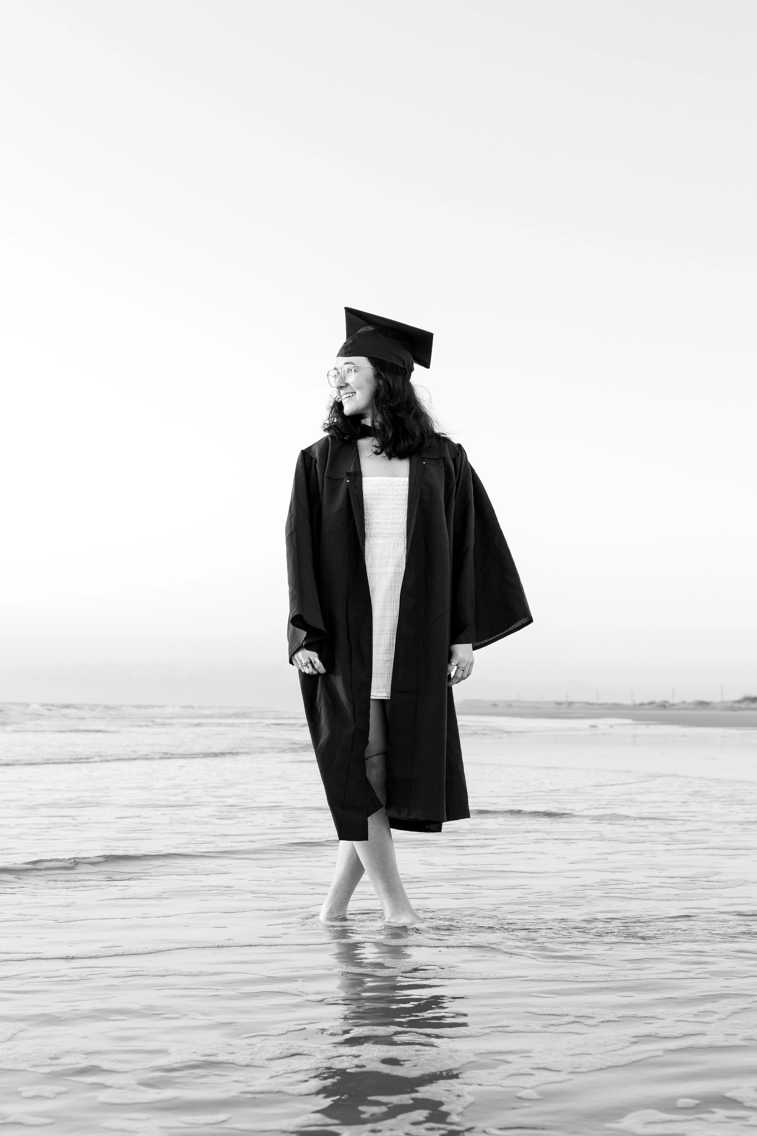 A woman wearing a graduation gown and cap, standing in shallow water on a beach, smiling and looking to her left.