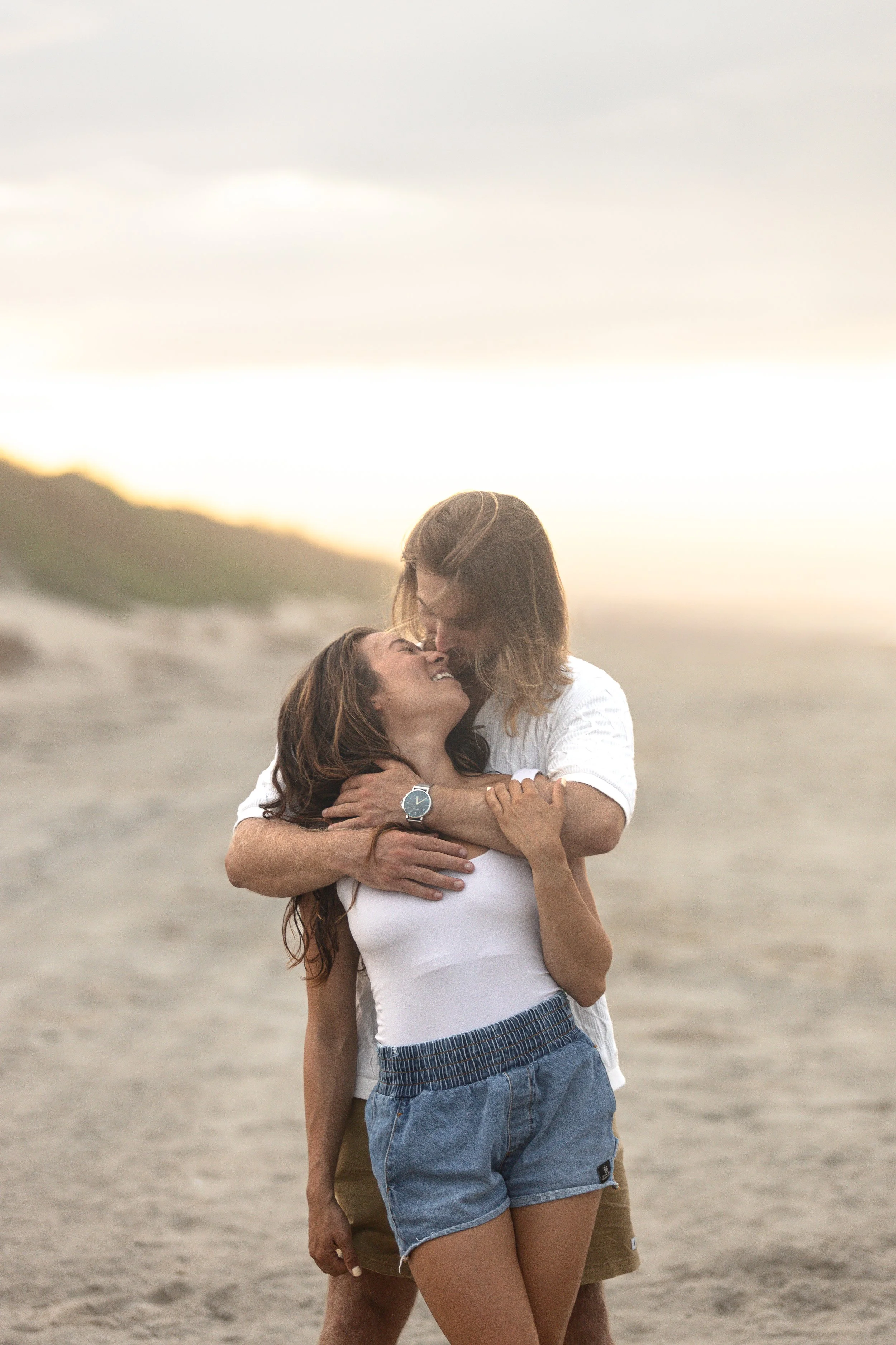 A couple embracing on a sandy beach during sunset, smiling and looking happy.