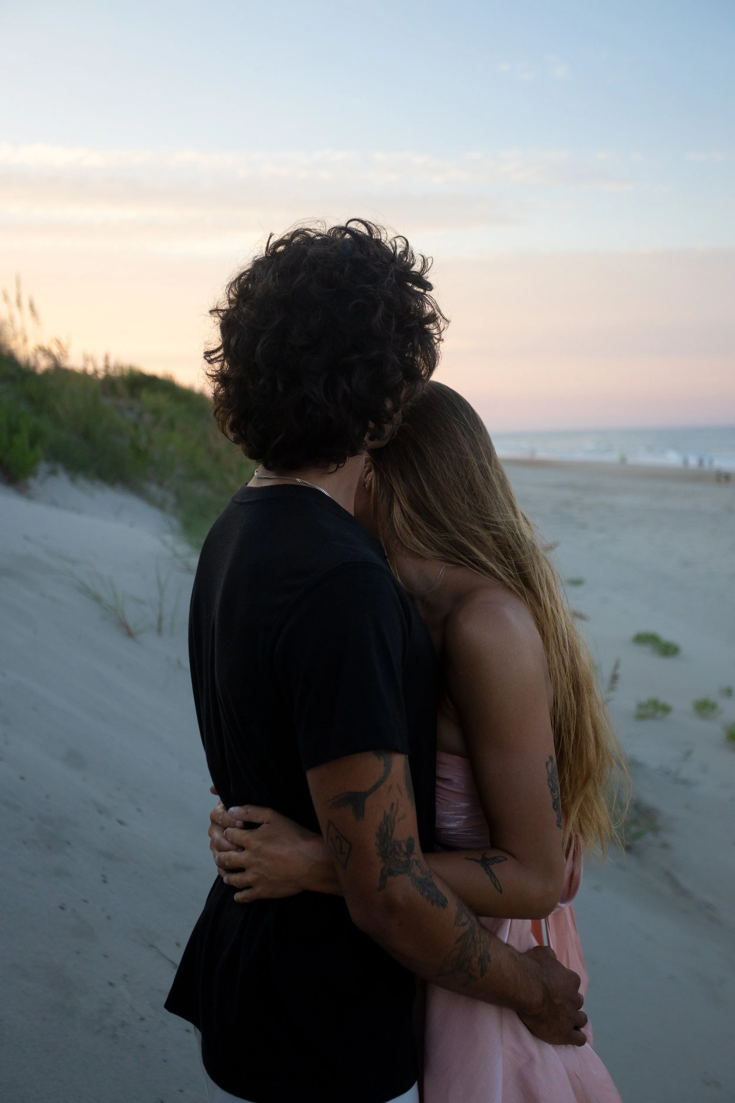A couple embraces on a beach at sunset, with their faces close, the man's hair curled, wearing black, and the woman's long hair down, wearing pink, with tattoos visible on their arms.