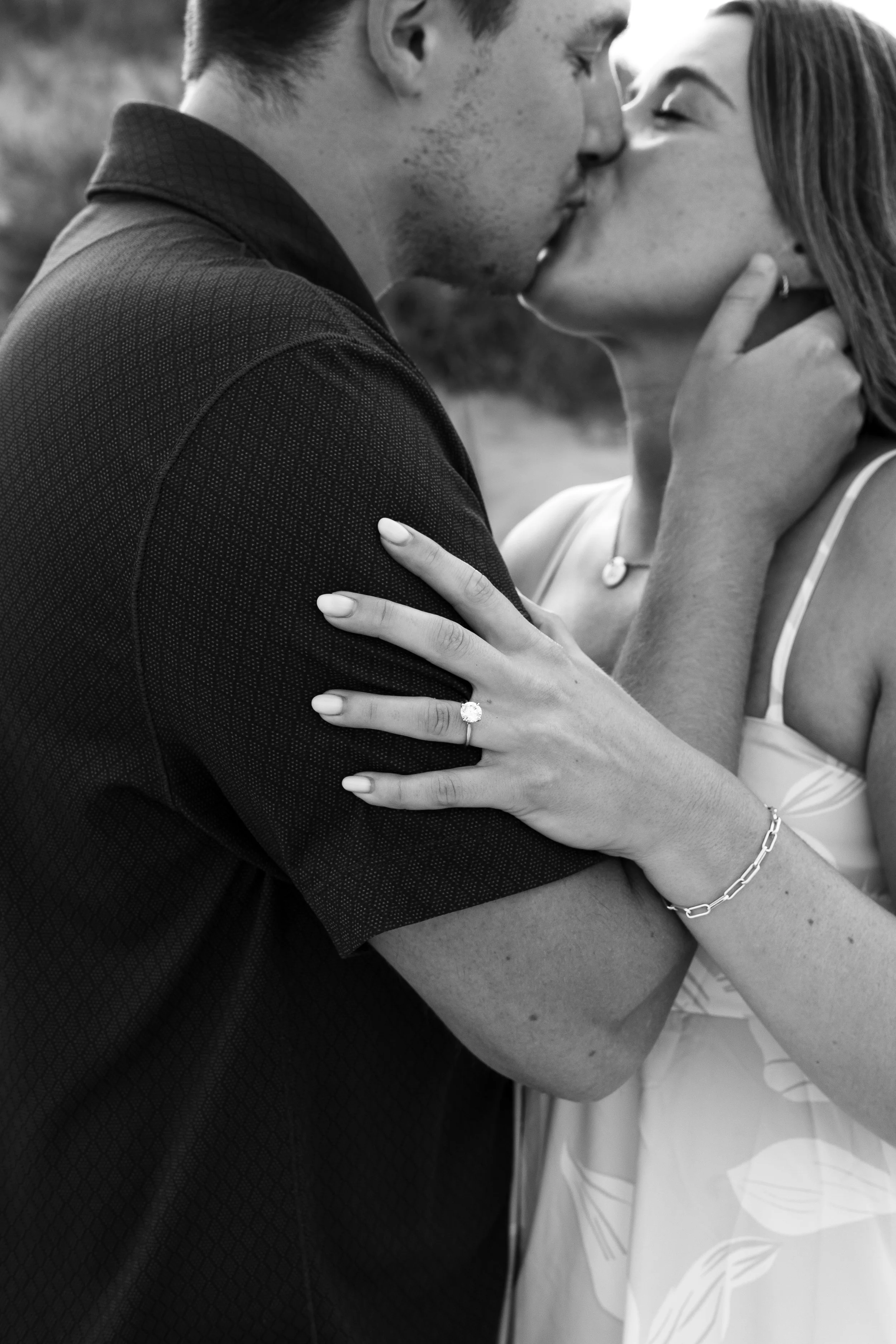 A black and white photo of a couple kissing, with the woman showing her engagement ring and wearing a bracelet, while the man holds her face.
