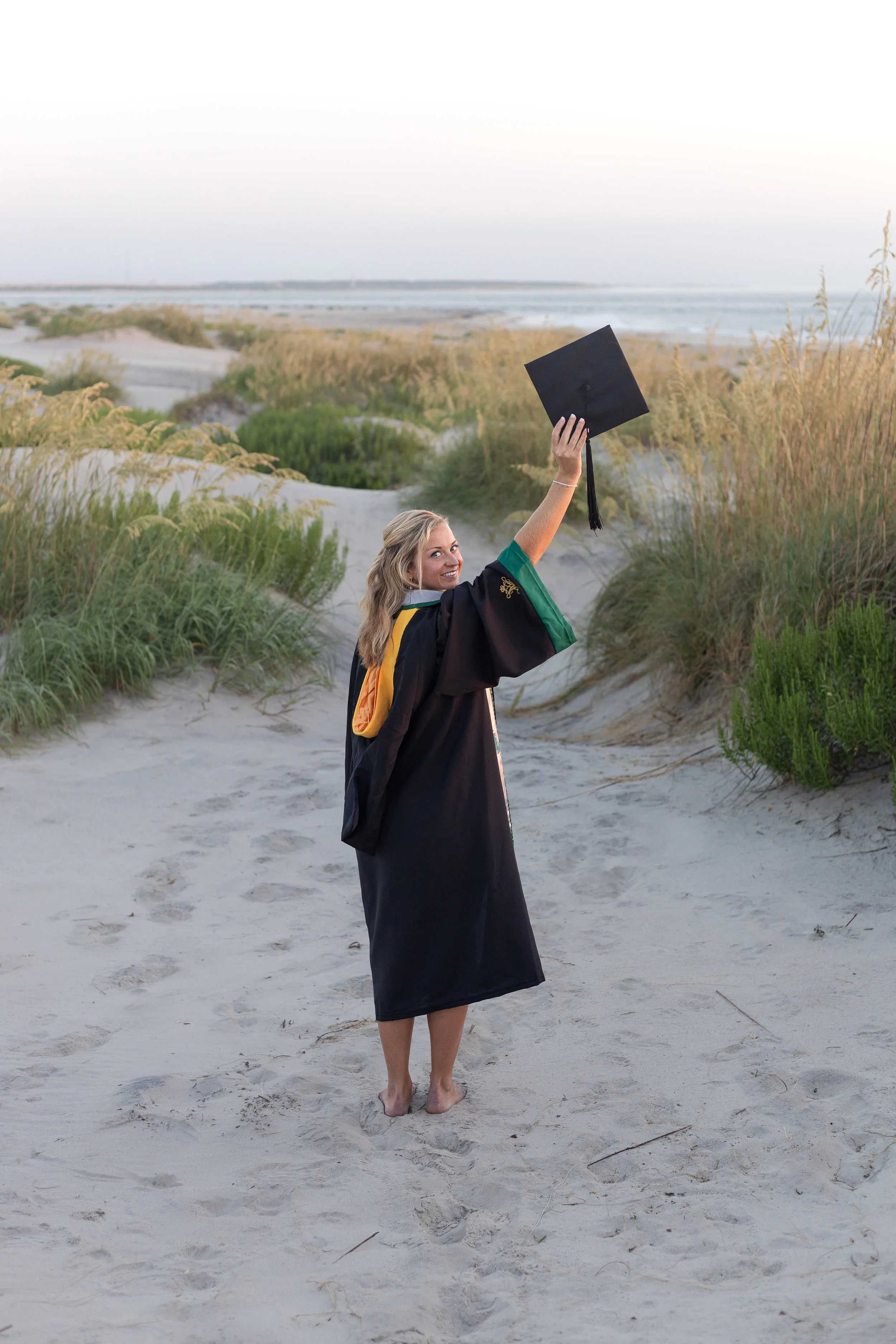 A young woman in a black graduation gown and cap standing barefoot on sandy dunes at the beach, holding her cap up in the air and smiling.