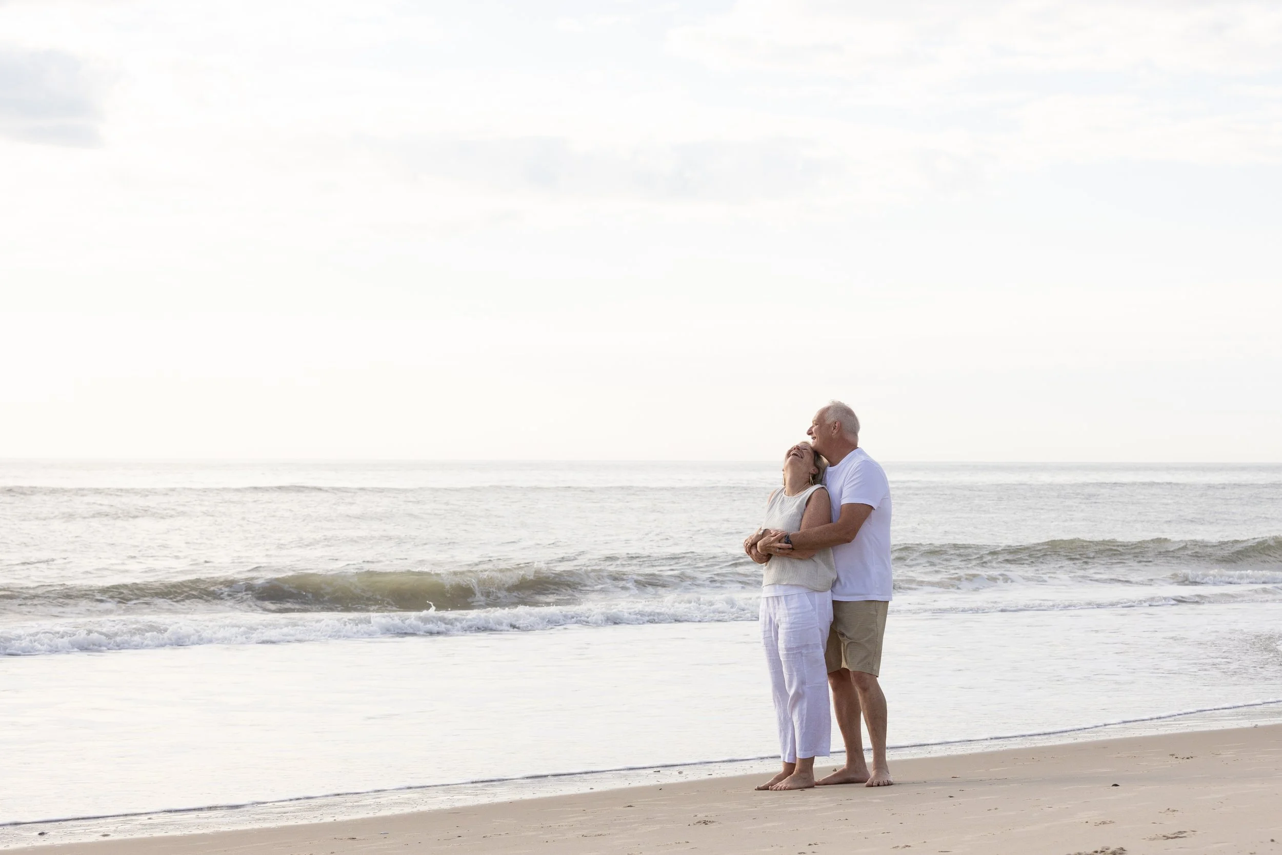 A loving couple embraces on the beach near the ocean, with waves crashing behind them and a partly cloudy sky above.
